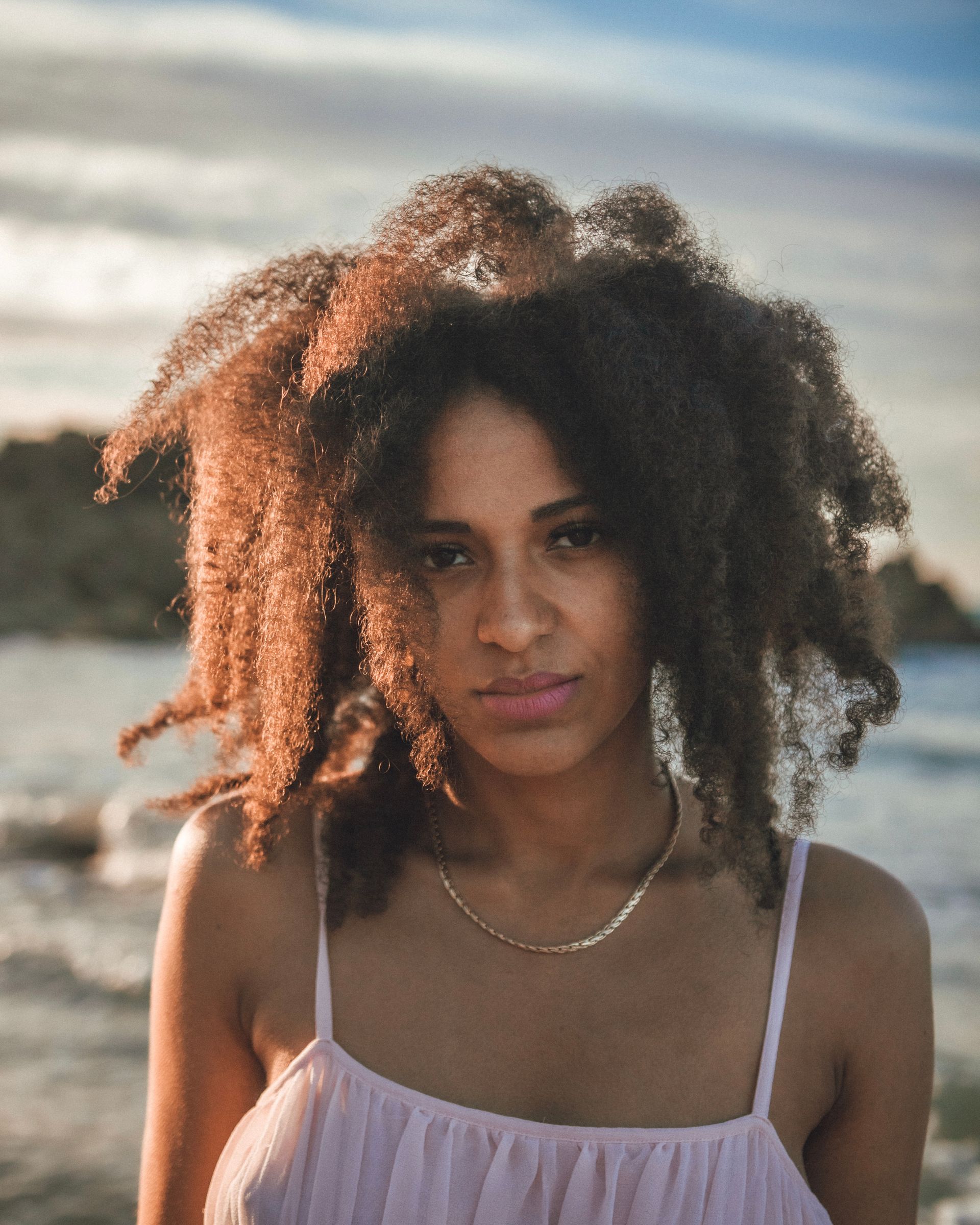 Femme aux cheveux bouclés dans une robe rose clair, regardant la caméra, arrière-plan de plage.