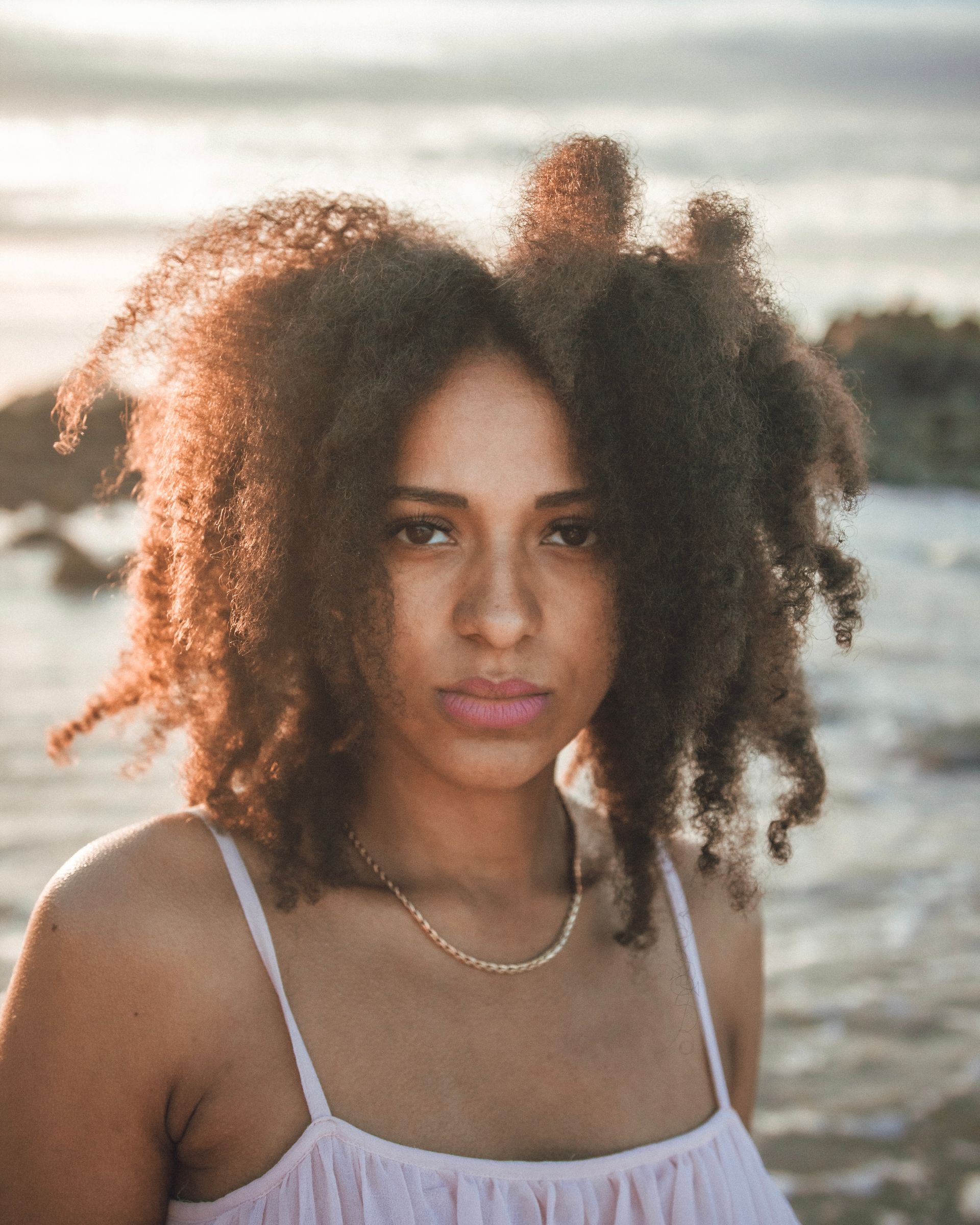 Femme aux cheveux bruns bouclés, dans un débardeur rose clair, regardant la caméra près de l'eau.