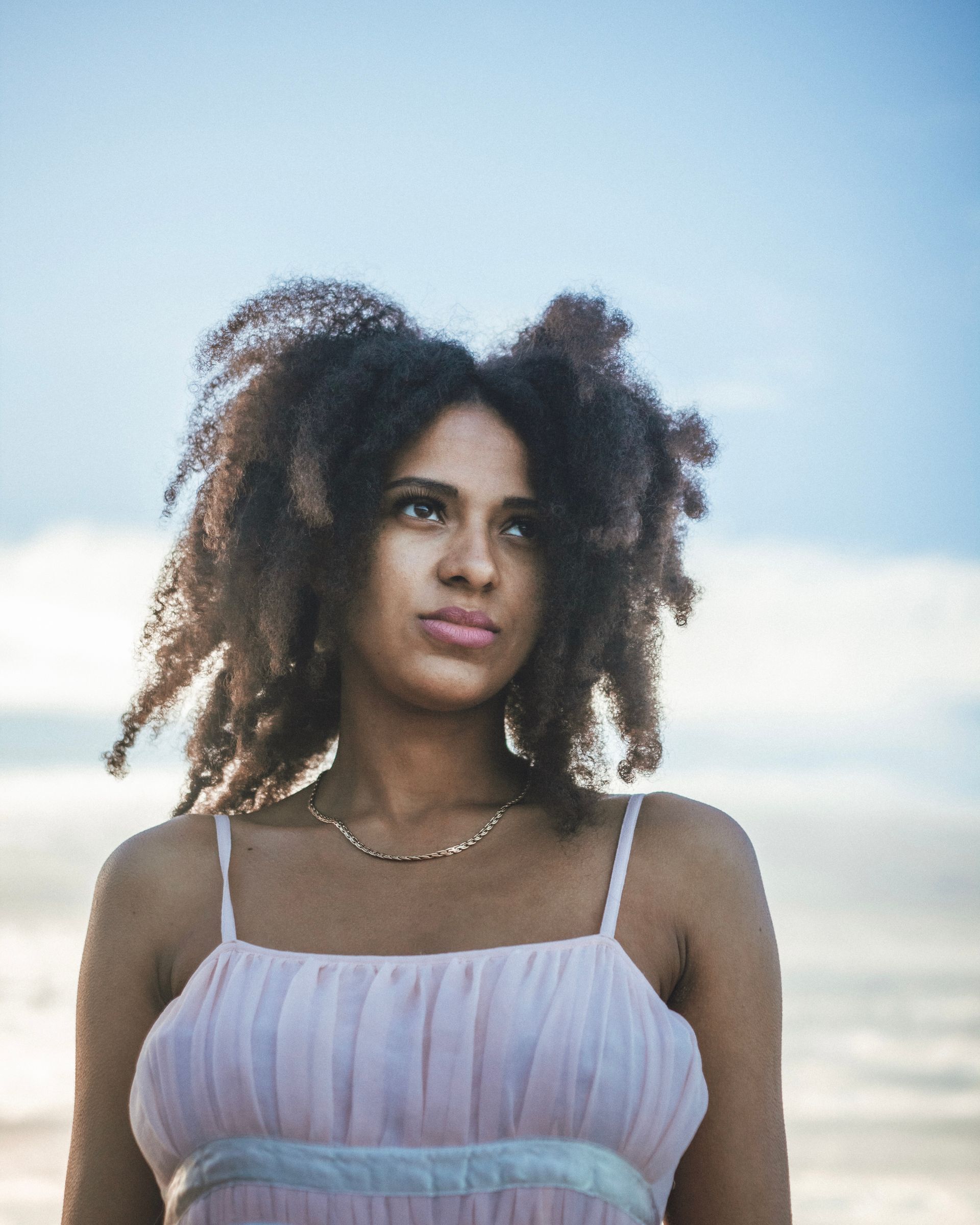 Femme aux cheveux bouclés portant une robe rose, regardant pensivement sur un fond de ciel bleu.