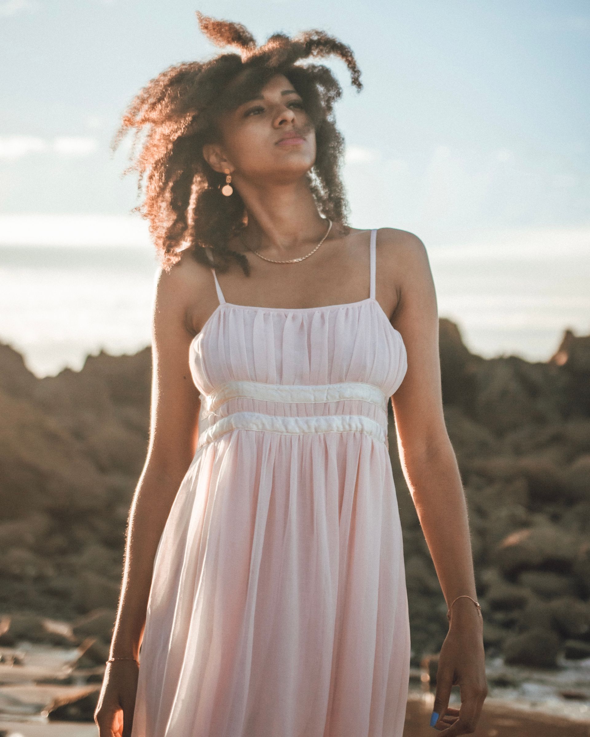 Femme en robe rose regardant le soleil, les cheveux au vent, sur une plage.