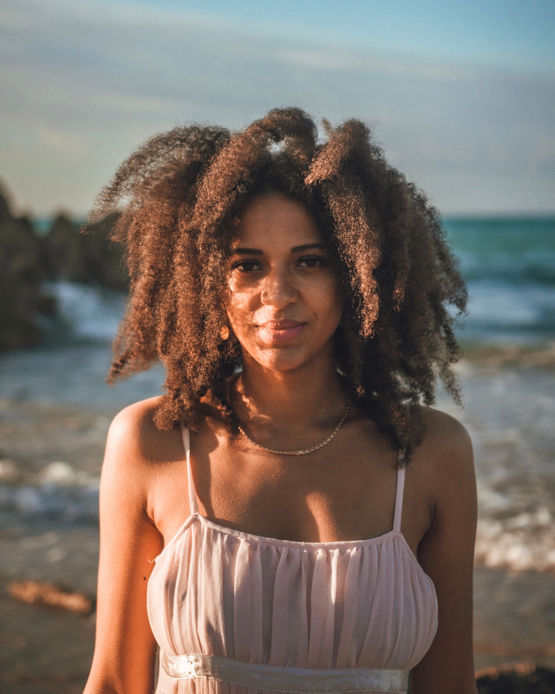 Une femme aux cheveux bruns bouclés dans une robe rose sourit à la caméra sur une plage.