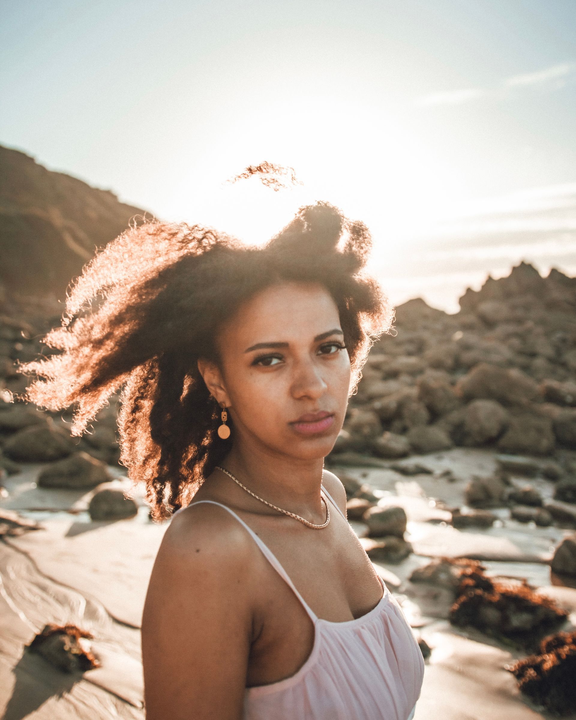 Femme aux cheveux bouclés dans une robe rose clair, sur une plage rocheuse au coucher du soleil.