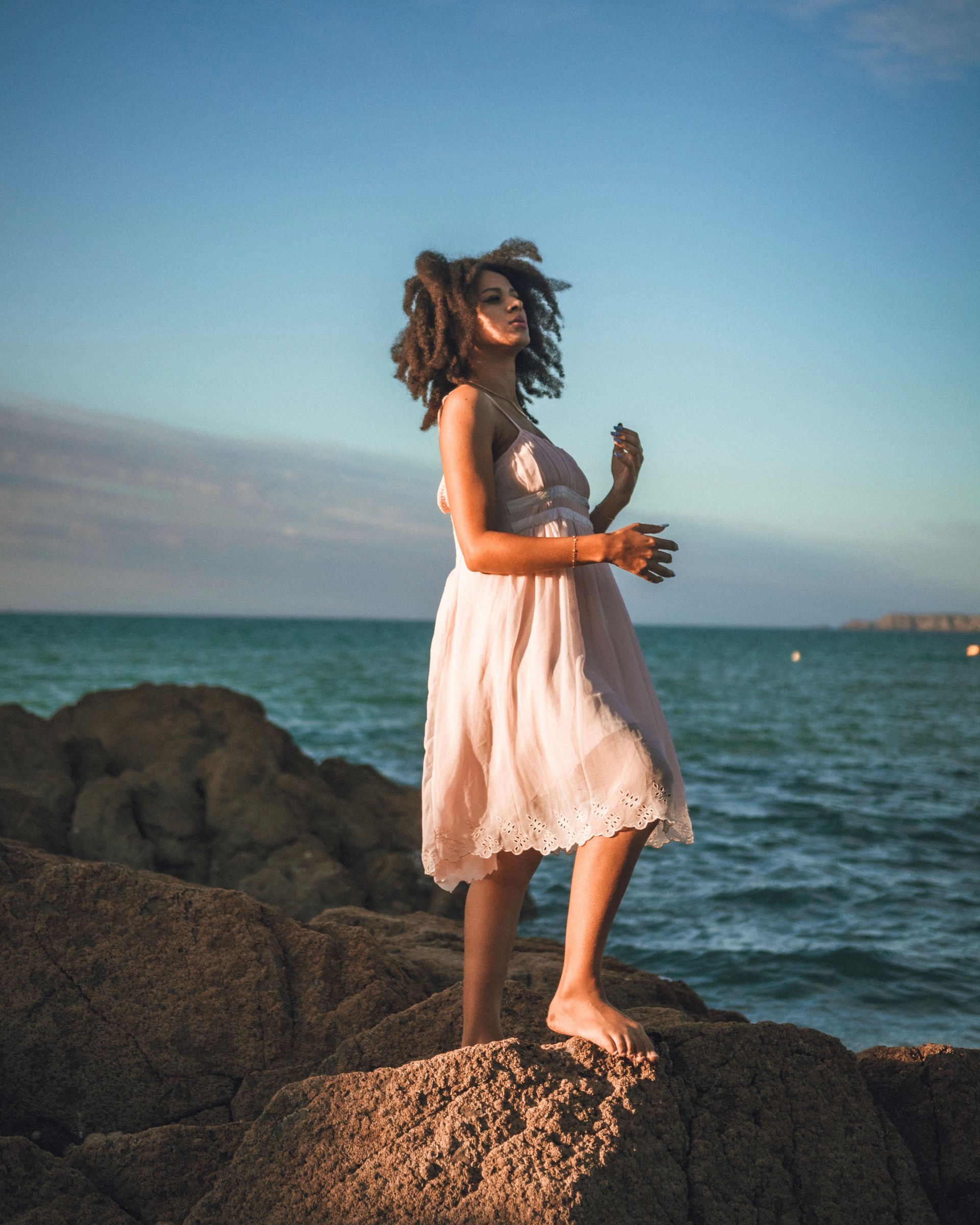 Une femme en robe rose se tient sur des rochers au bord de la mer, regardant vers le spectateur, lumière dorée.