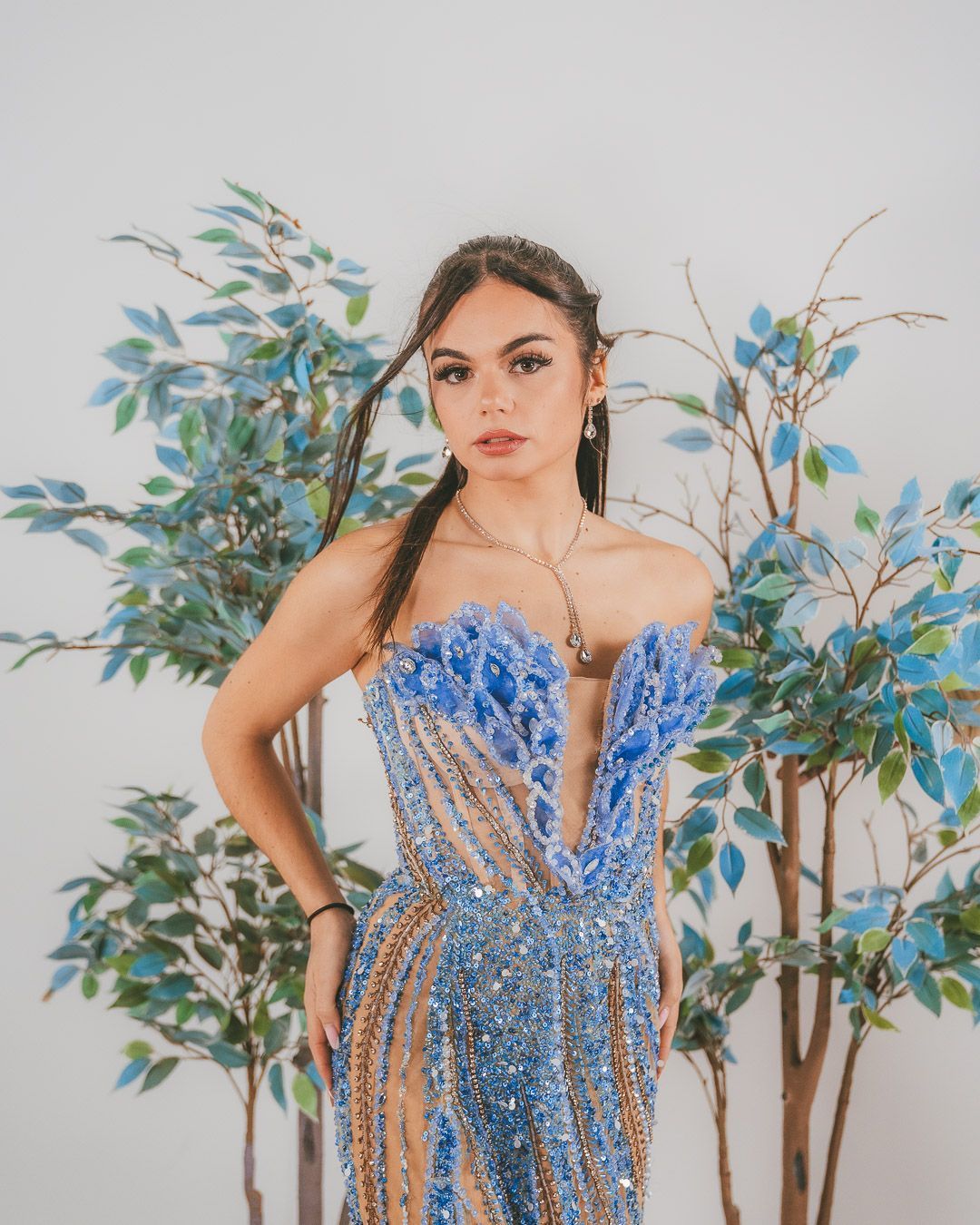 Une femme en robe perlée bleue pose devant des branches feuillues.