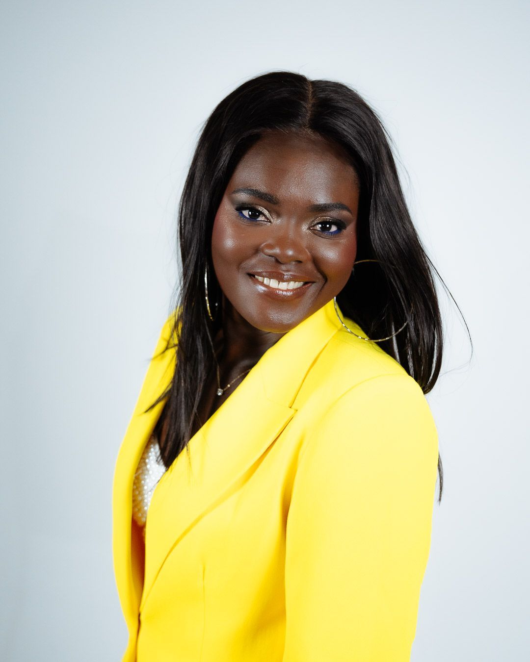 Une jeune femme à la peau foncée sourit, vêtue d'un blazer jaune vif, d'un haut blanc ; prise de vue en studio.