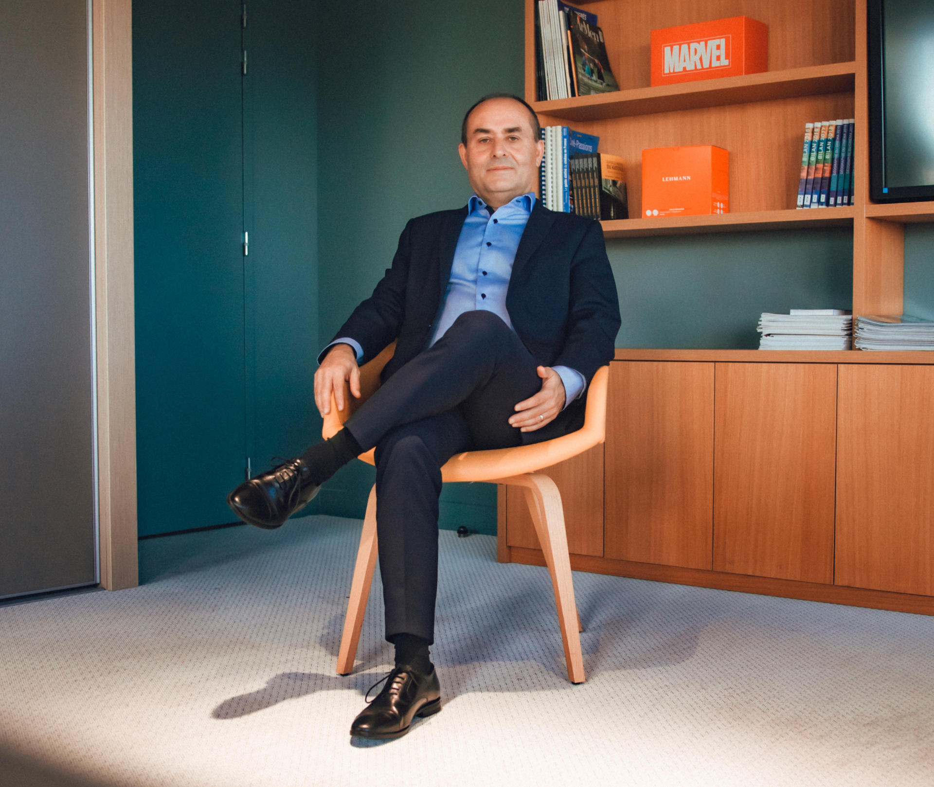 Un homme en costume est assis les jambes croisées dans un bureau moderne, avec une étagère en arrière-plan.
