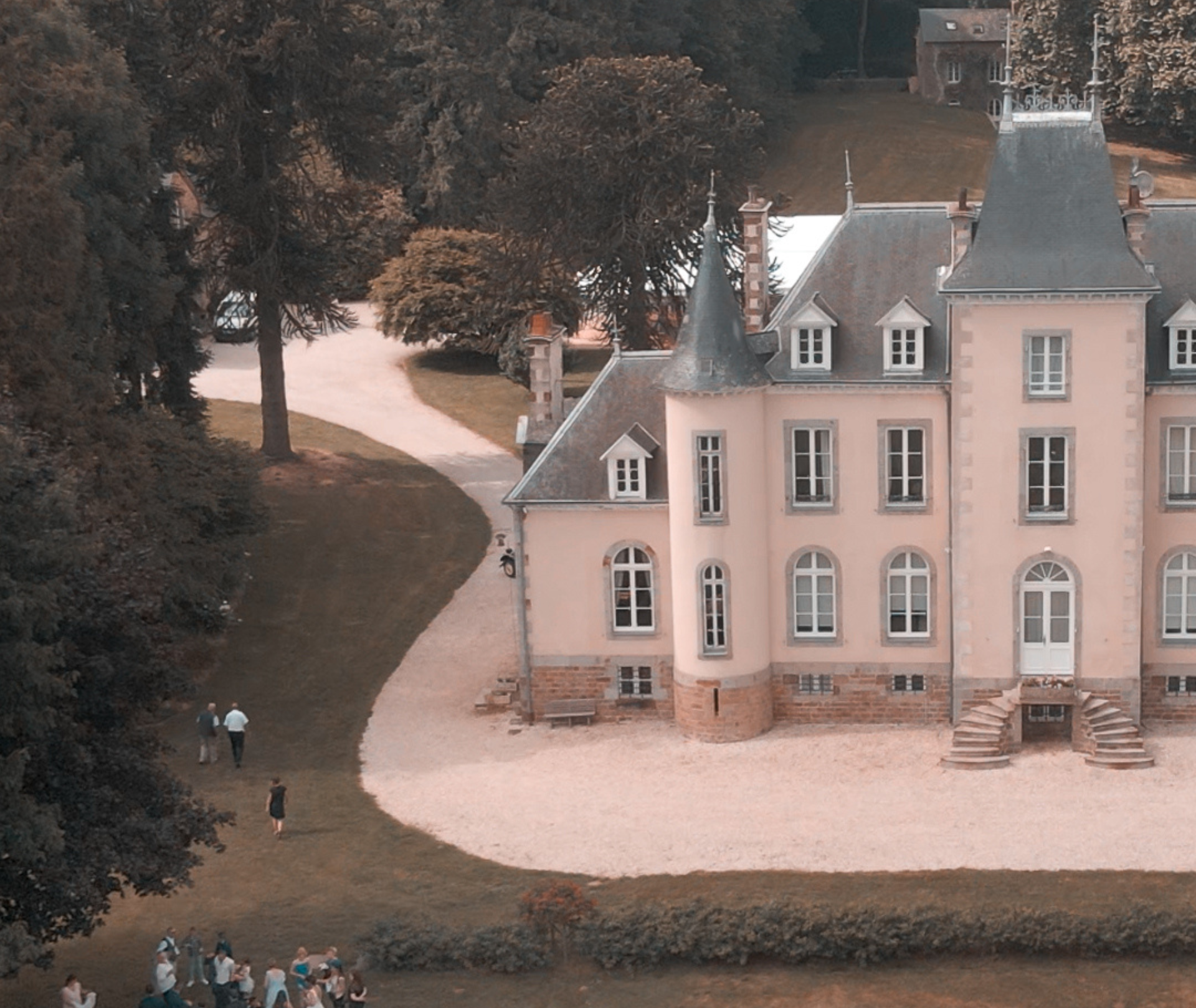 Château rose avec tourelles et allée sinueuse, des gens marchant sur la pelouse.