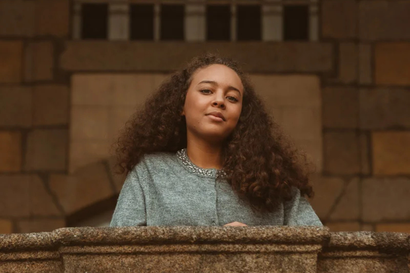 Une jeune femme aux cheveux bouclés sourit, appuyée sur un balcon en pierre. Vêtue d'un pull gris, elle est dehors.