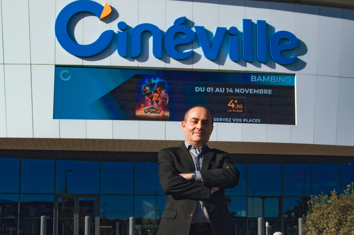 Homme devant un cinéma Cinéville, bras croisés. Panneau bleu avec logo et affiche du film.