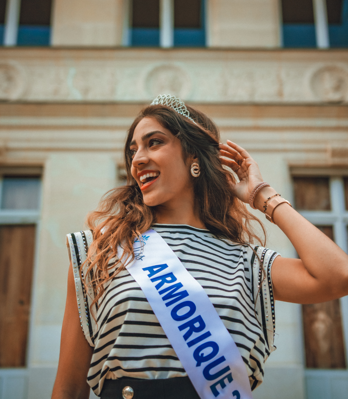 Femme portant un diadème et une ceinture sur laquelle est écrit « Armorique » souriant devant un bâtiment.