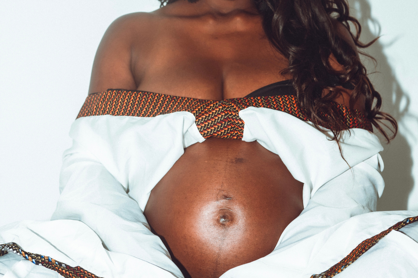 Femme noire enceinte au ventre nu, portant un haut à motifs blanc et marron, posée devant un fond blanc.