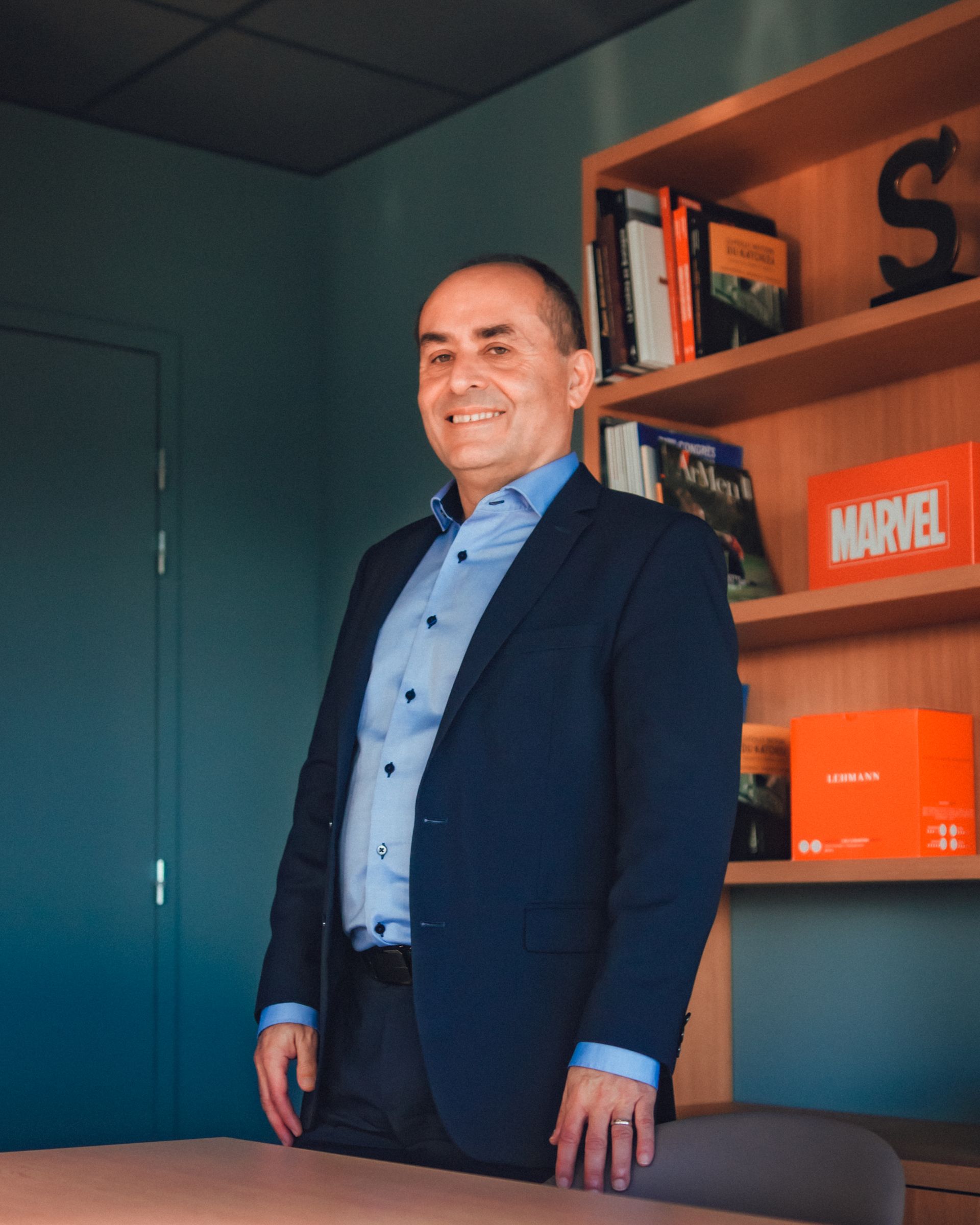Un homme en costume souriant, appuyé contre son bureau. Une bibliothèque en arrière-plan.