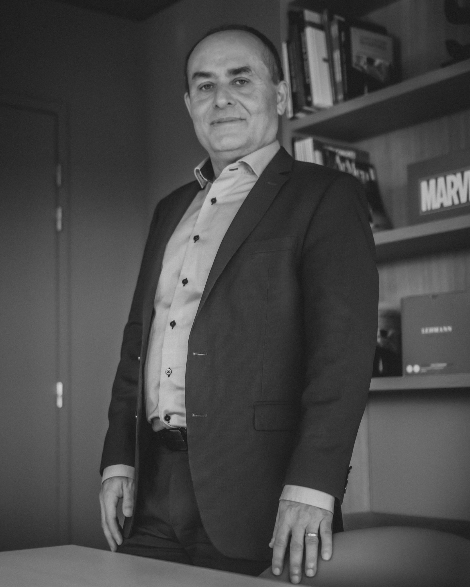 Homme en costume, debout dans un bureau. Bibliothèque en arrière-plan, panneau « Marvel » visible.