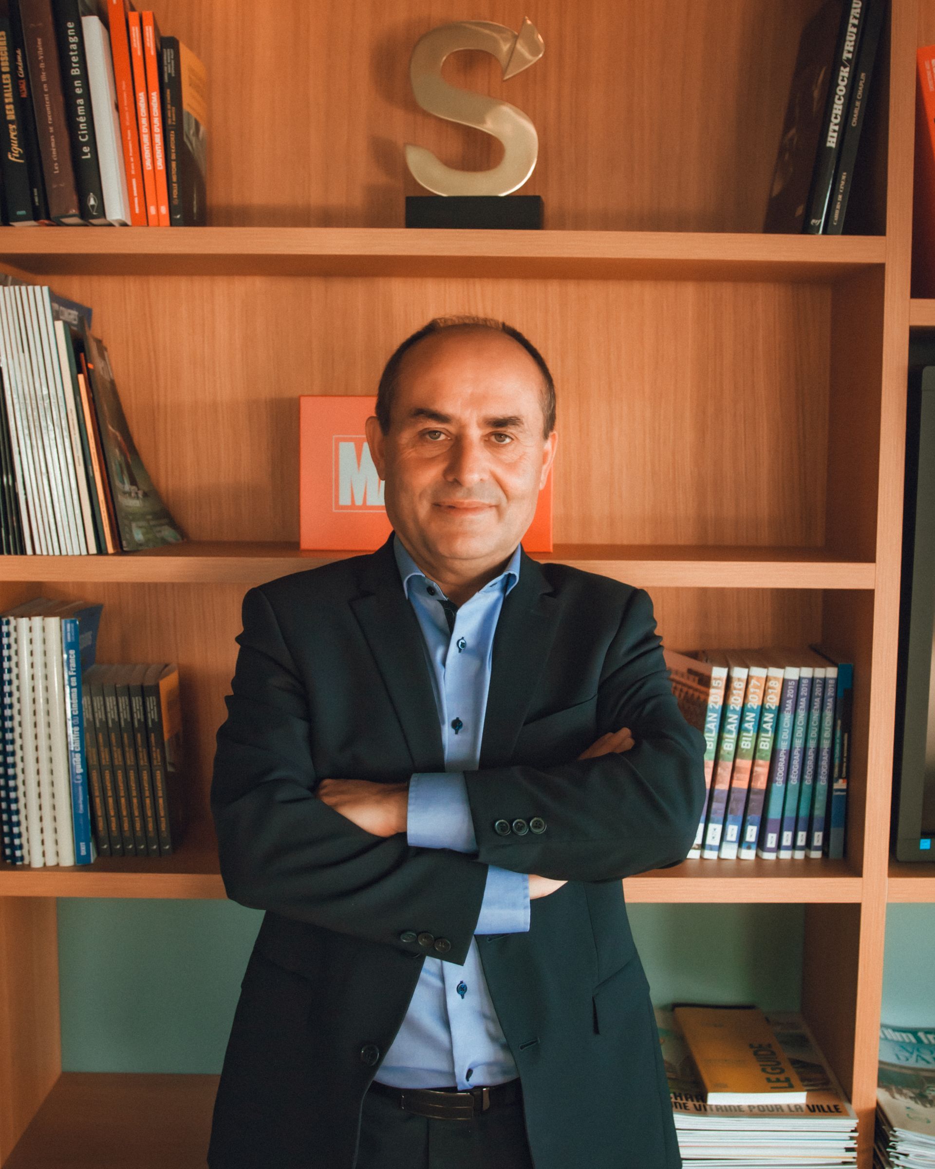 Homme en costume, les bras croisés, debout devant une bibliothèque.