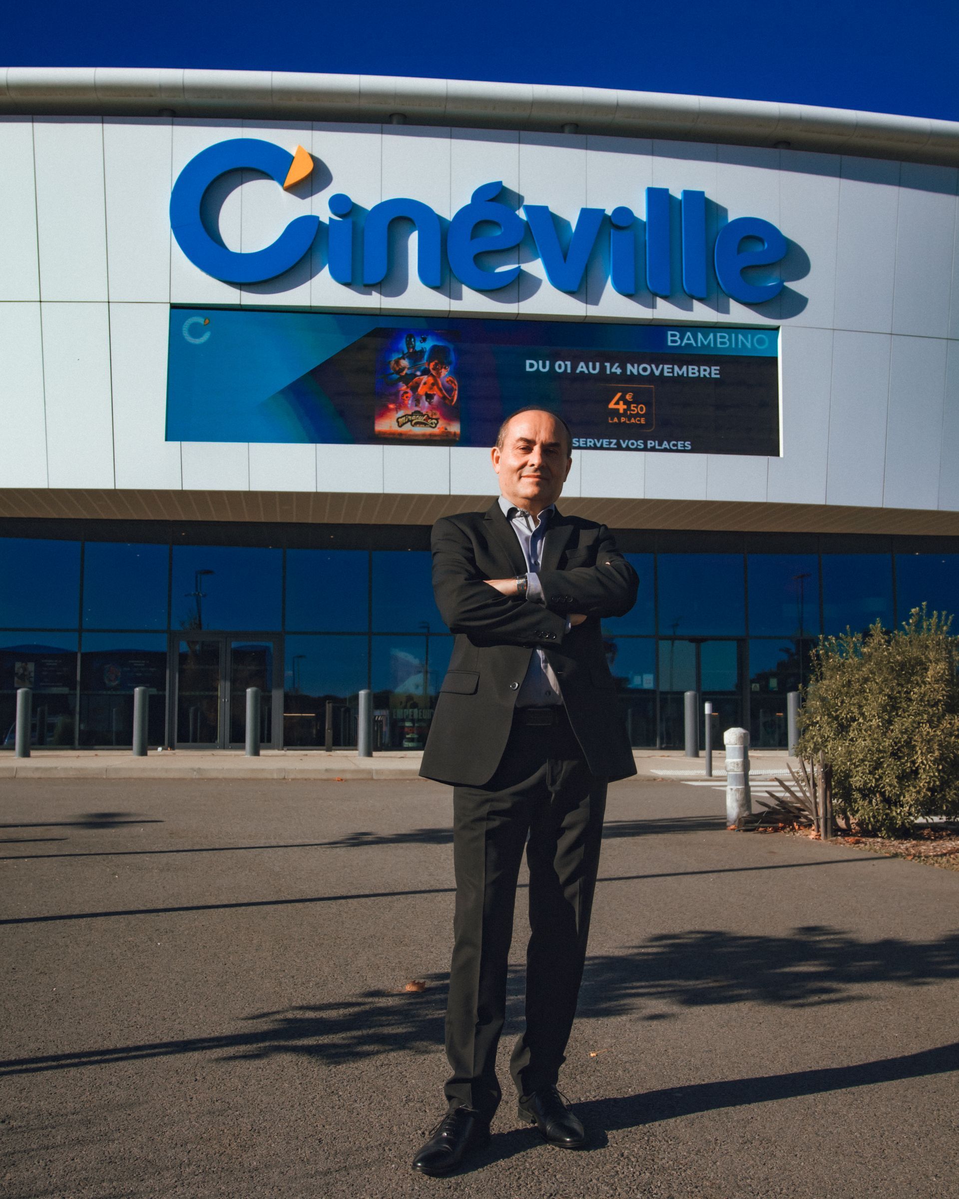Homme en costume debout devant un cinéma Cineville, bras croisés, ciel bleu.