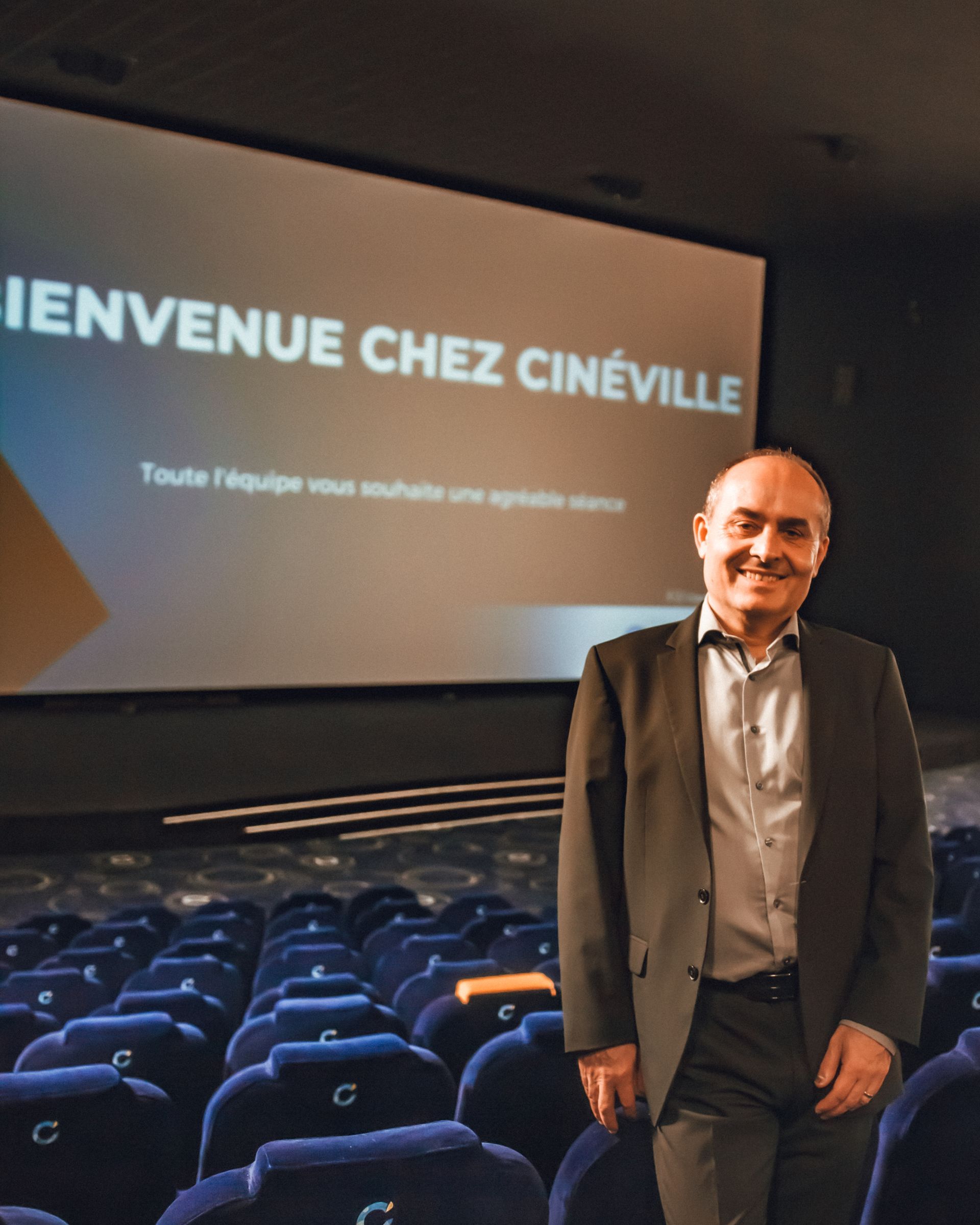 Un homme en costume sourit dans un cinéma Cineville. « Bienvenue chez Cineville » à l'écran, sièges bleus.
