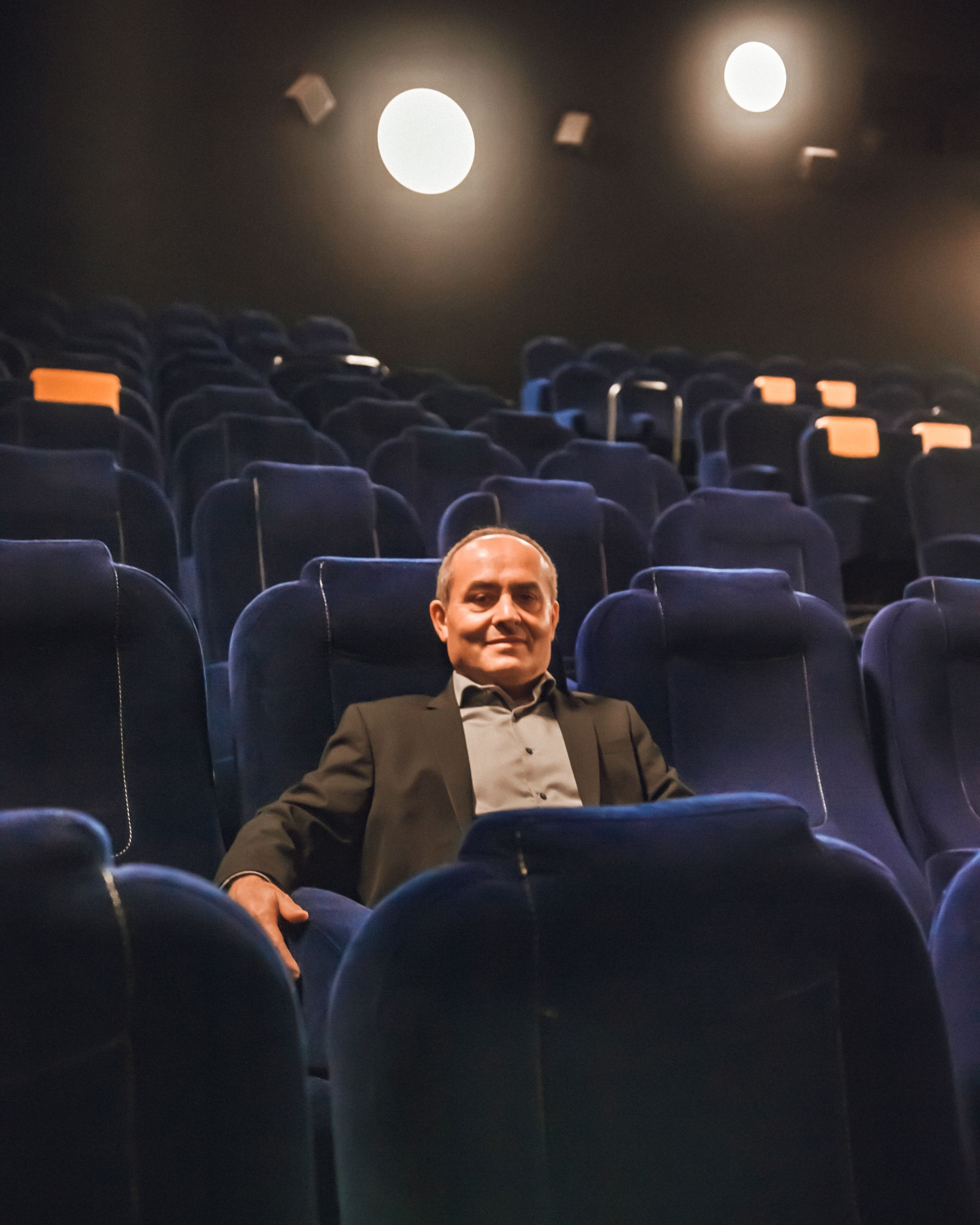 Un homme est assis et sourit dans une salle de cinéma sombre avec des sièges bleus vides et des lumières au plafond.