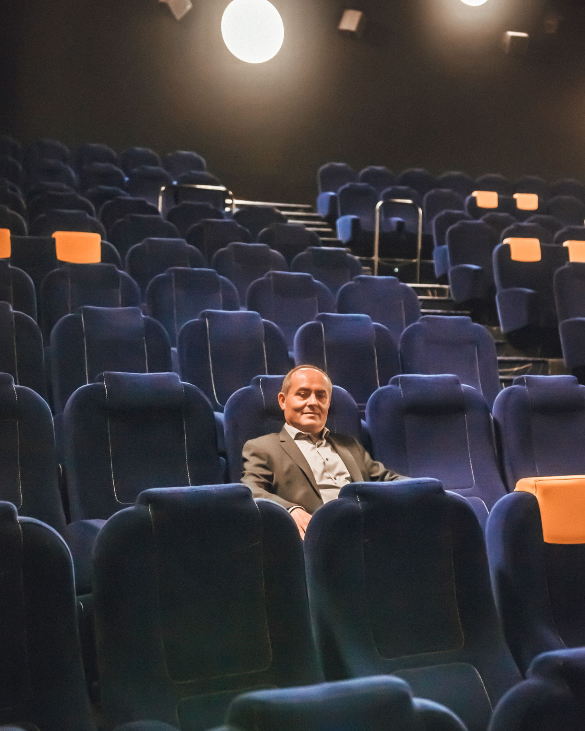 Un homme en costume est assis sur un siège bleu dans une salle de cinéma.