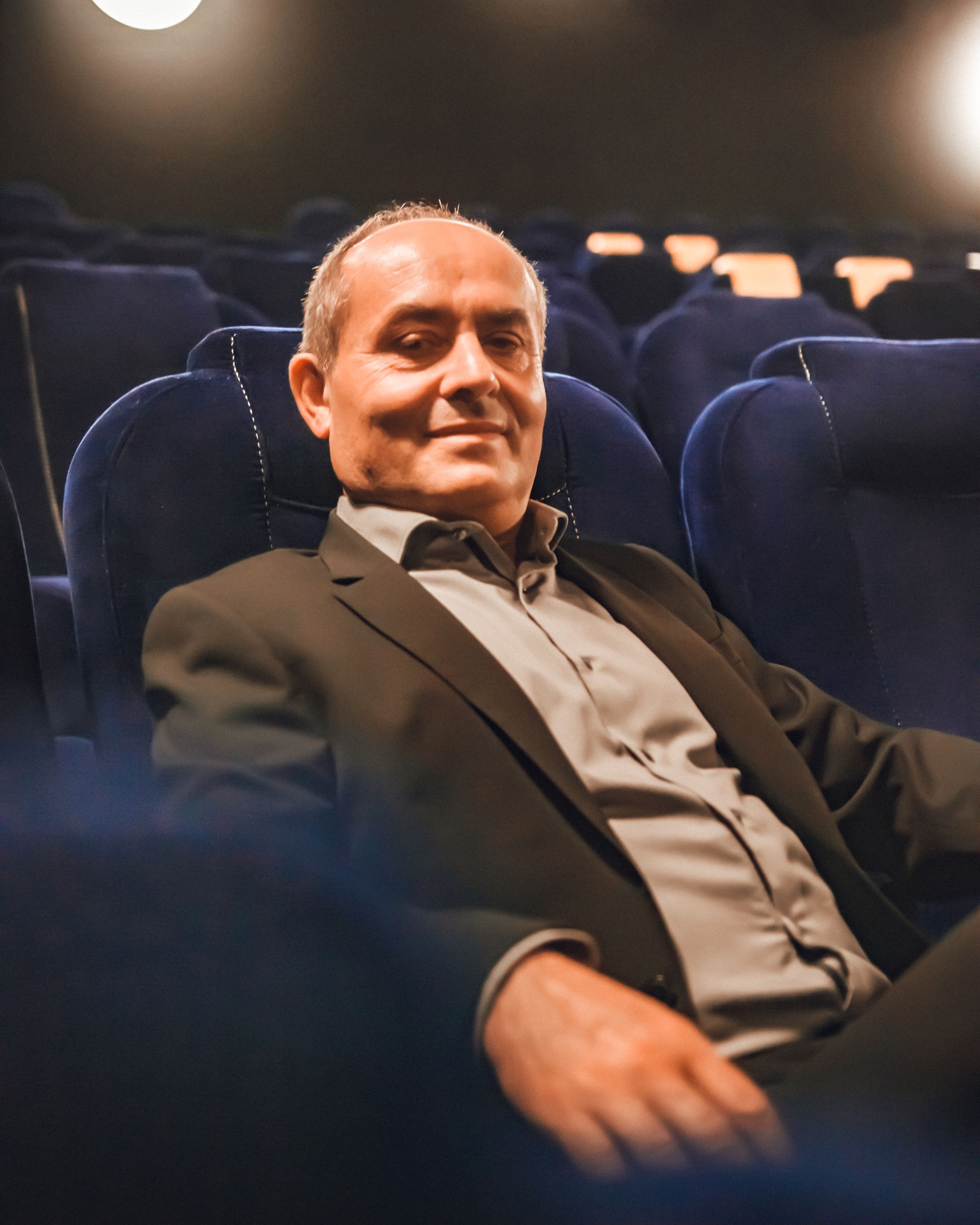 Un homme en costume, assis dans un fauteuil de cinéma, souriant, regardant droit devant. Sièges bleus, lumière tamisée.