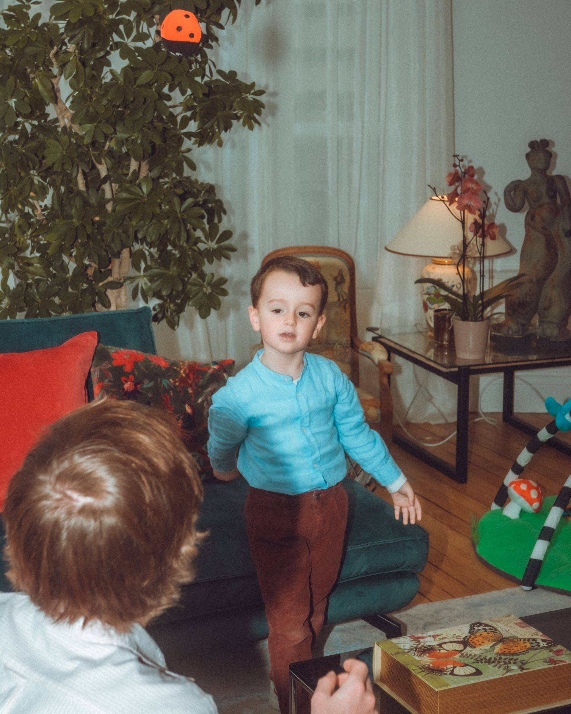 Enfant en pull bleu et pantalon marron, debout dans un salon, face à la caméra. Une autre personne est partiellement visible.