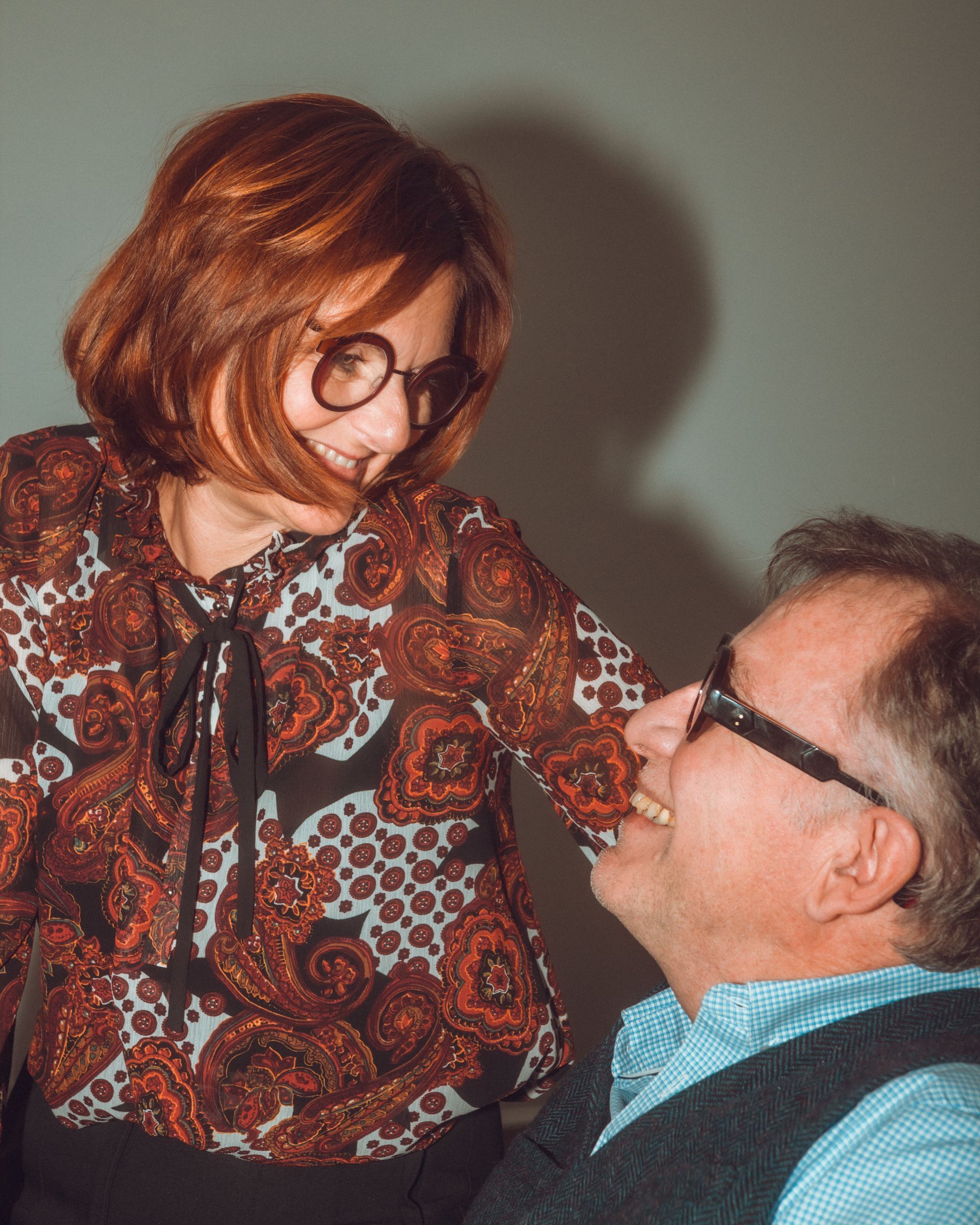 Une femme aux cheveux roux sourit à un homme ; tous deux portent des lunettes et sourient, dans une pièce au mur gris.