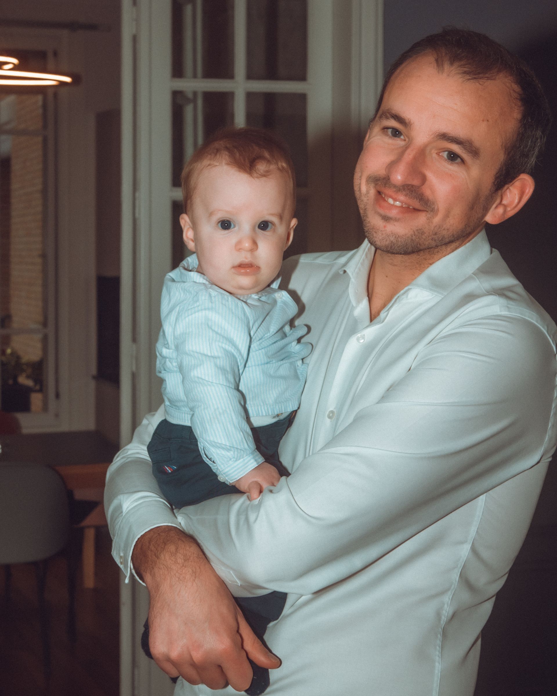 Un homme souriant tient un bébé dans ses bras dans une pièce lumineuse. Tous deux portent des chemises blanches.