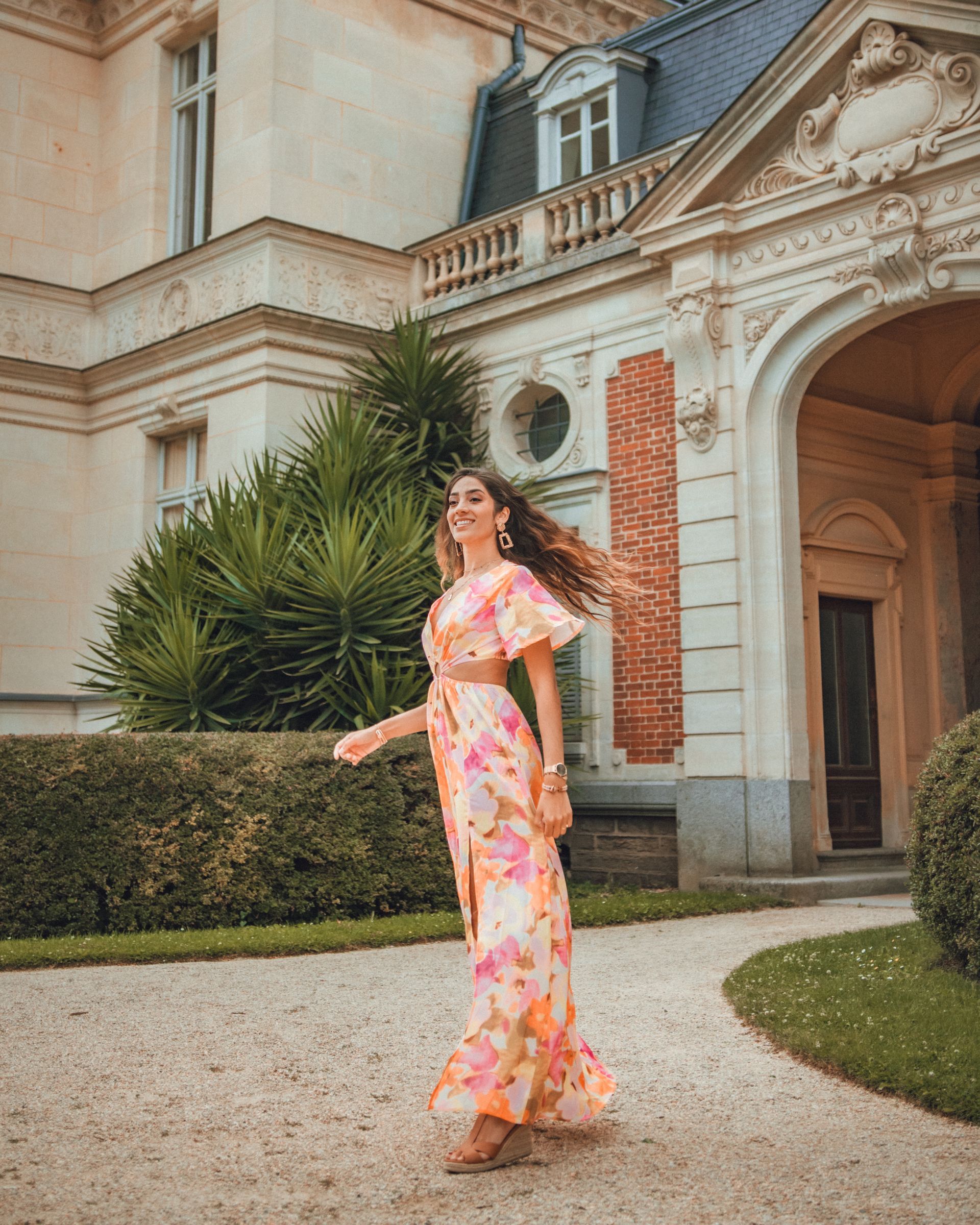 Une femme en robe fleurie virevolte devant un bâtiment majestueux avec un aménagement paysager soigné.