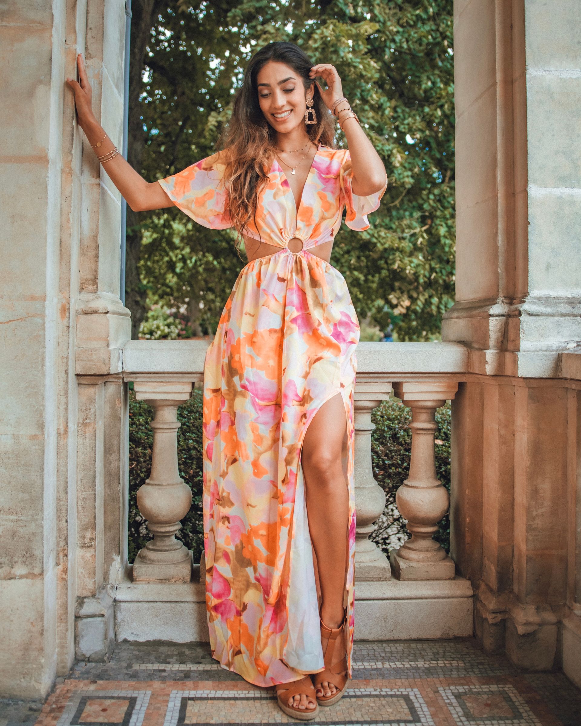 Une femme en robe longue fleurie pose sur un balcon, souriante, avec une fente haute sur la jambe.