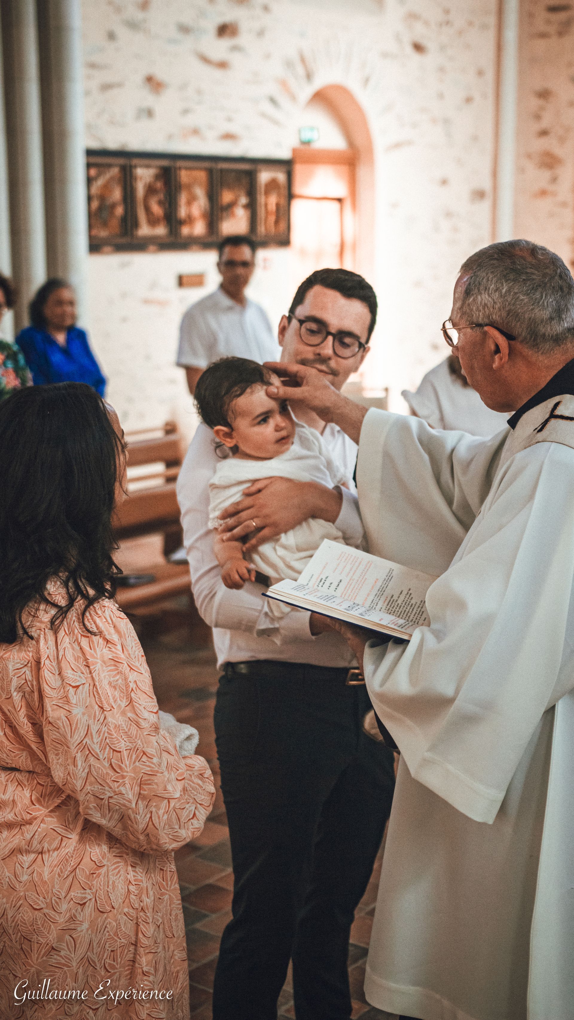 Un homme tient un bébé lors d'une cérémonie de baptême ; un prêtre oint le front du bébé dans une église.