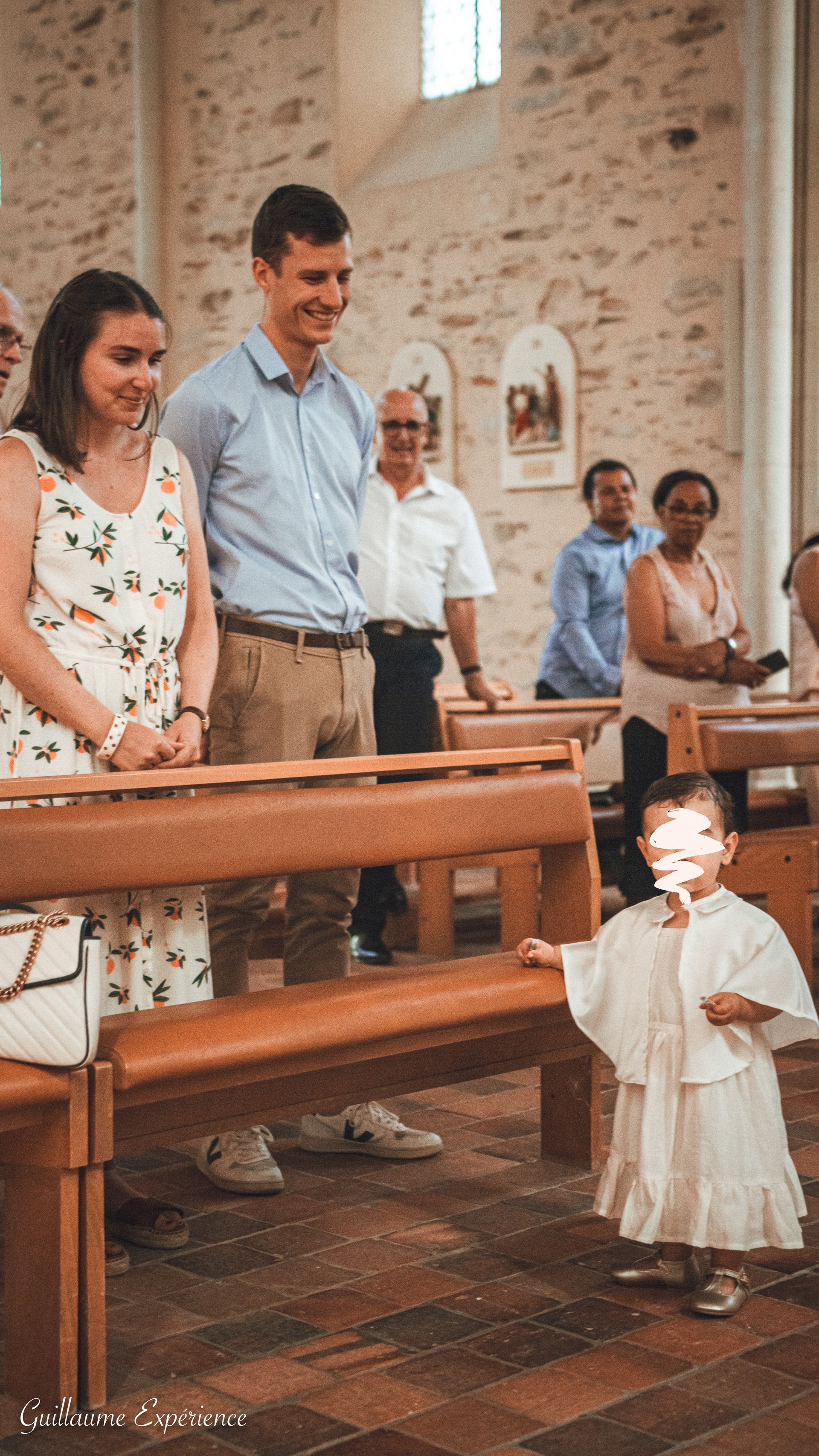 Un enfant en robe blanche de baptême dans une église. Parents et invités sourient.