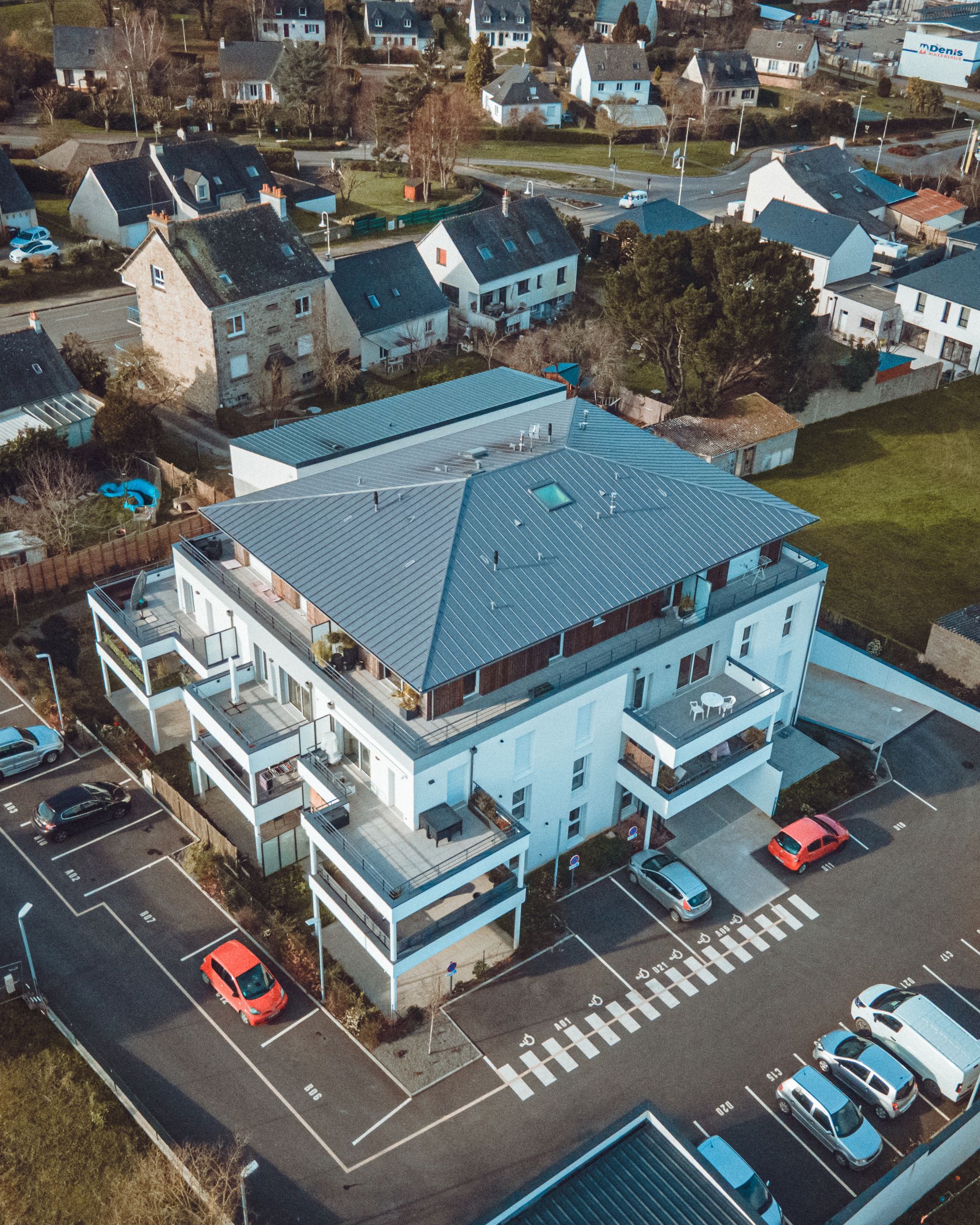 Vue aérienne d'un immeuble d'appartements moderne avec un toit bleu, des voitures garées et un quartier résidentiel.