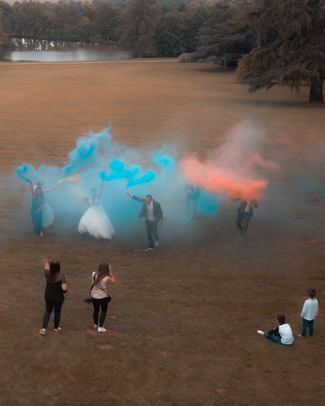 Cérémonie de mariage avec fumigènes colorés dans un champ ; mariée, marié, invités, photographes. Fumée bleue et orange.