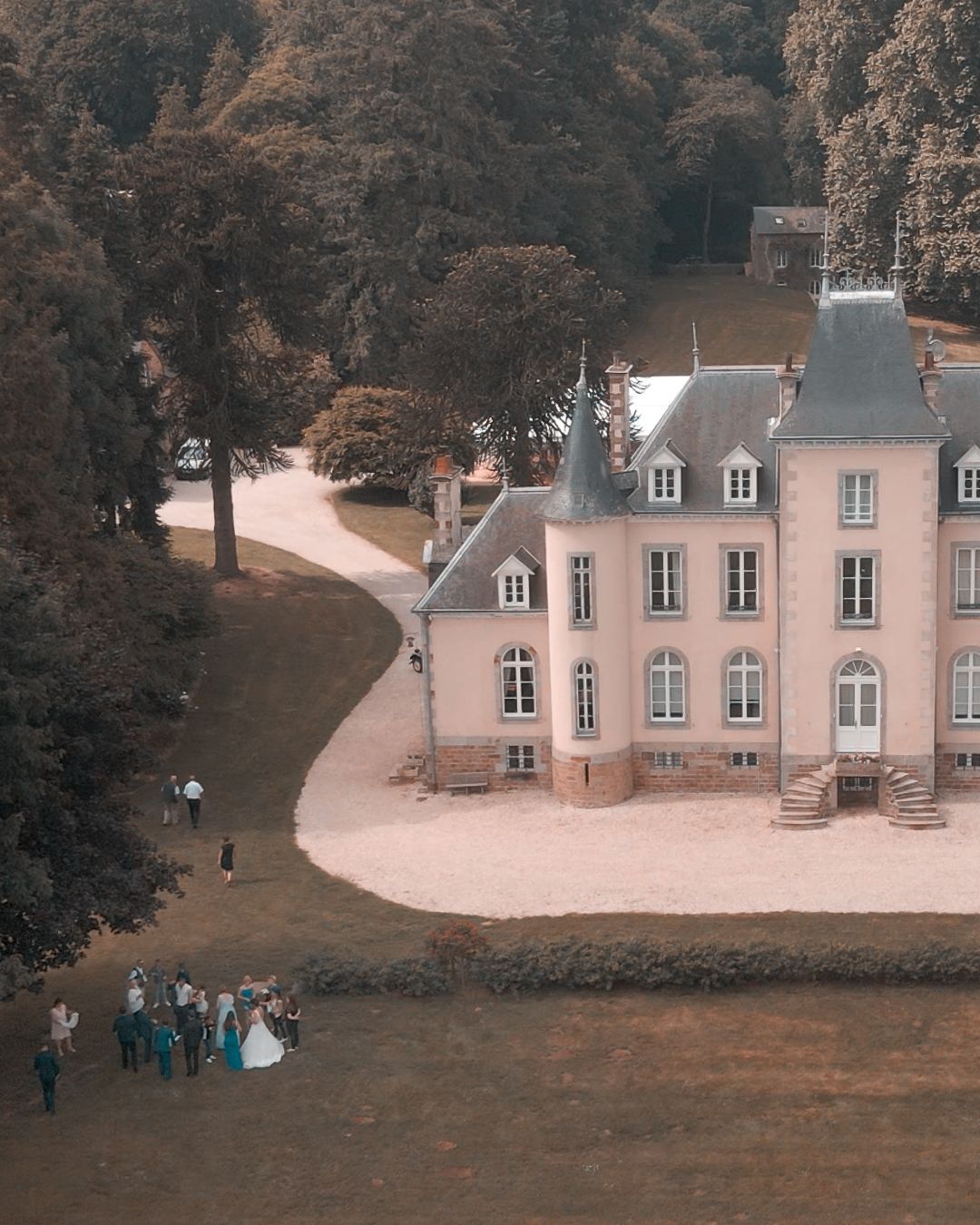 Fête de mariage devant un château. Bâtiment rose clair, allée sinueuse, arbres.