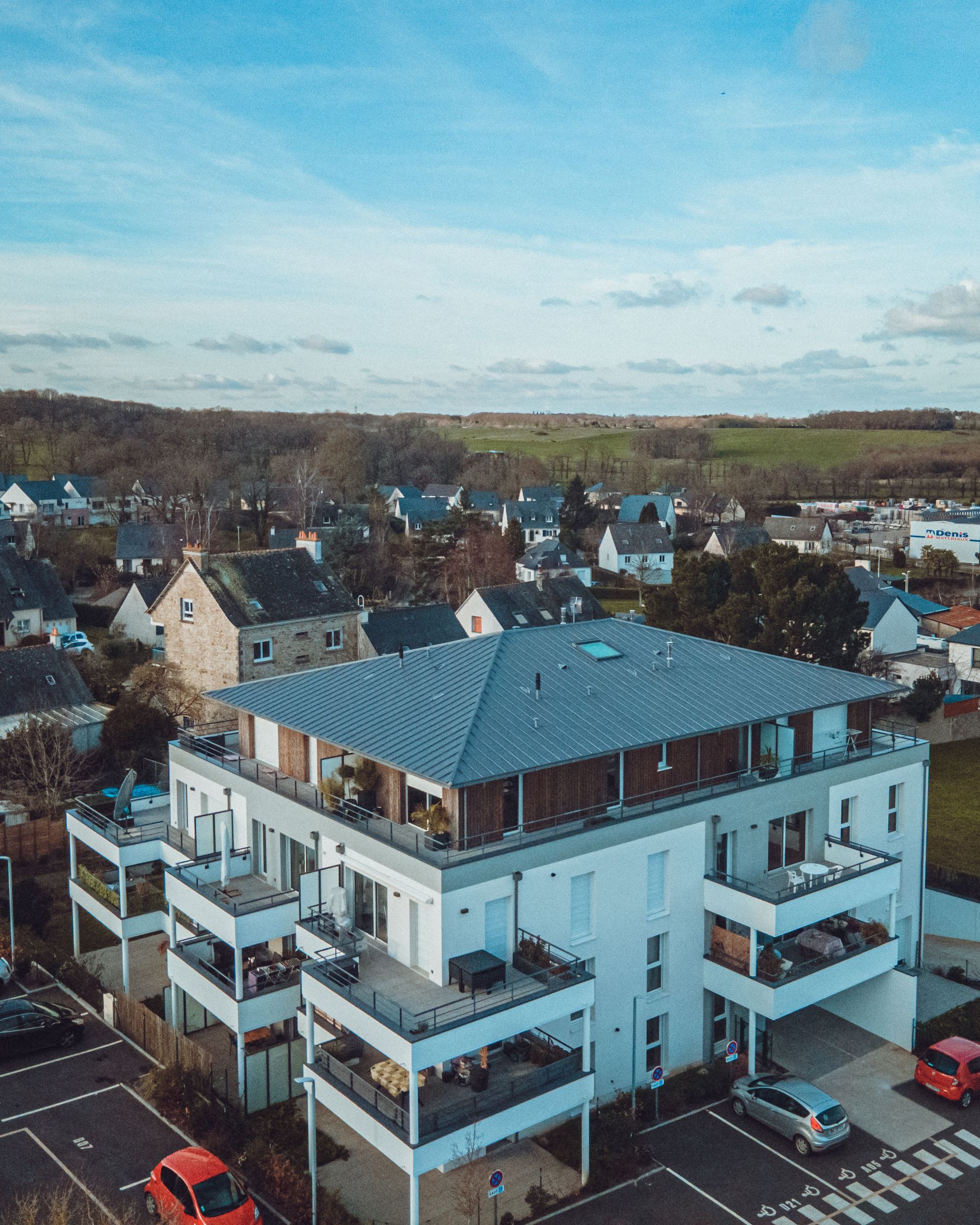 Immeuble d'appartements blanc avec balcons et parking, situé dans un décor de banlieue.