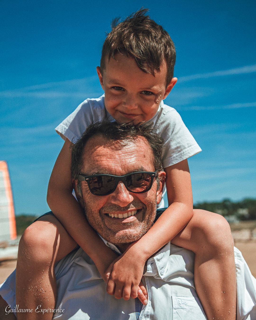 Un homme portant des lunettes de soleil sourit, un enfant assis sur ses épaules. Ciel bleu et plage en arrière-plan.