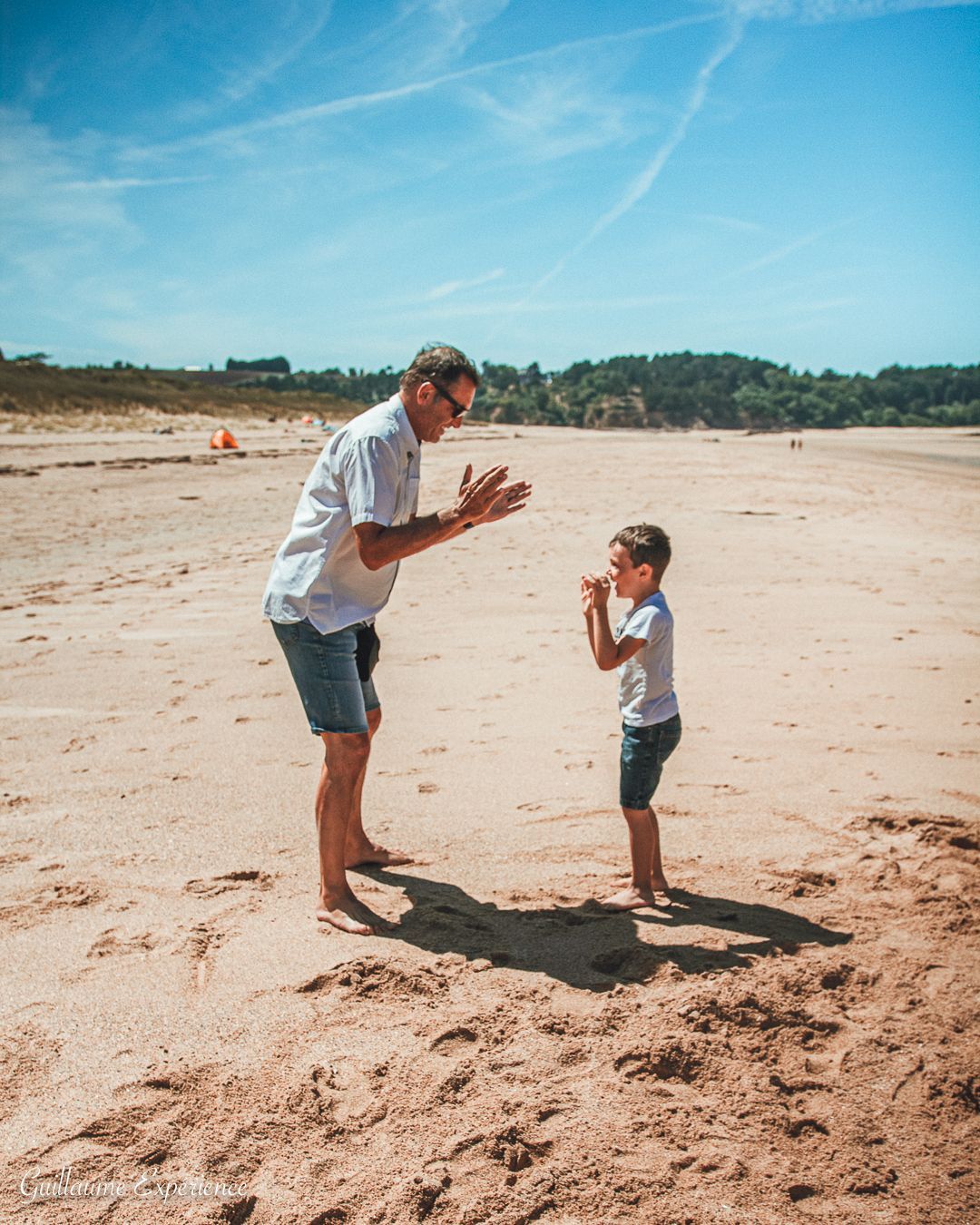 Un homme et un enfant sur une plage de sable, applaudissant. Un ciel bleu éclatant au-dessus de leurs têtes.