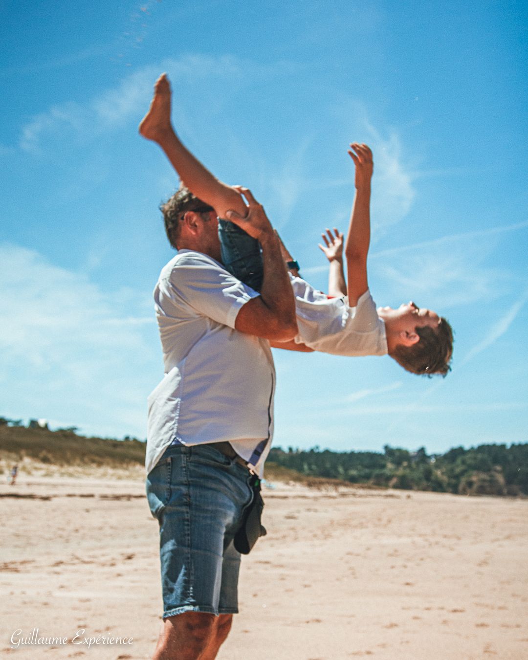 Un homme tenant un enfant la tête en bas sur une plage de sable sous un ciel bleu vif.