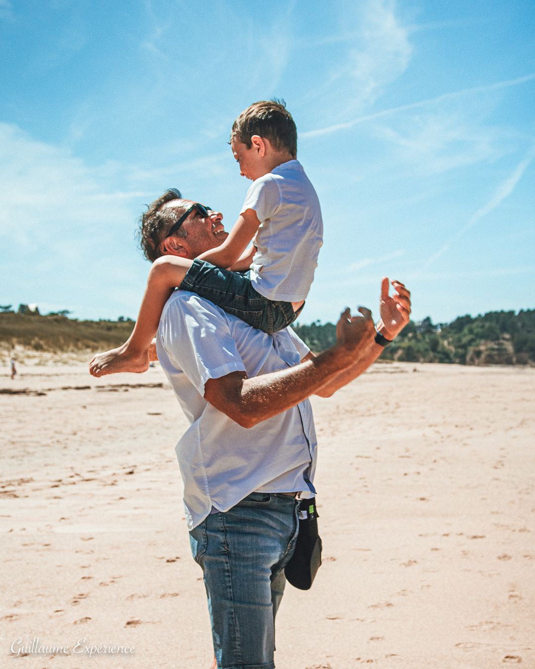 Un père porte son fils sur ses épaules à la plage ; tous deux sourient par une journée ensoleillée.
