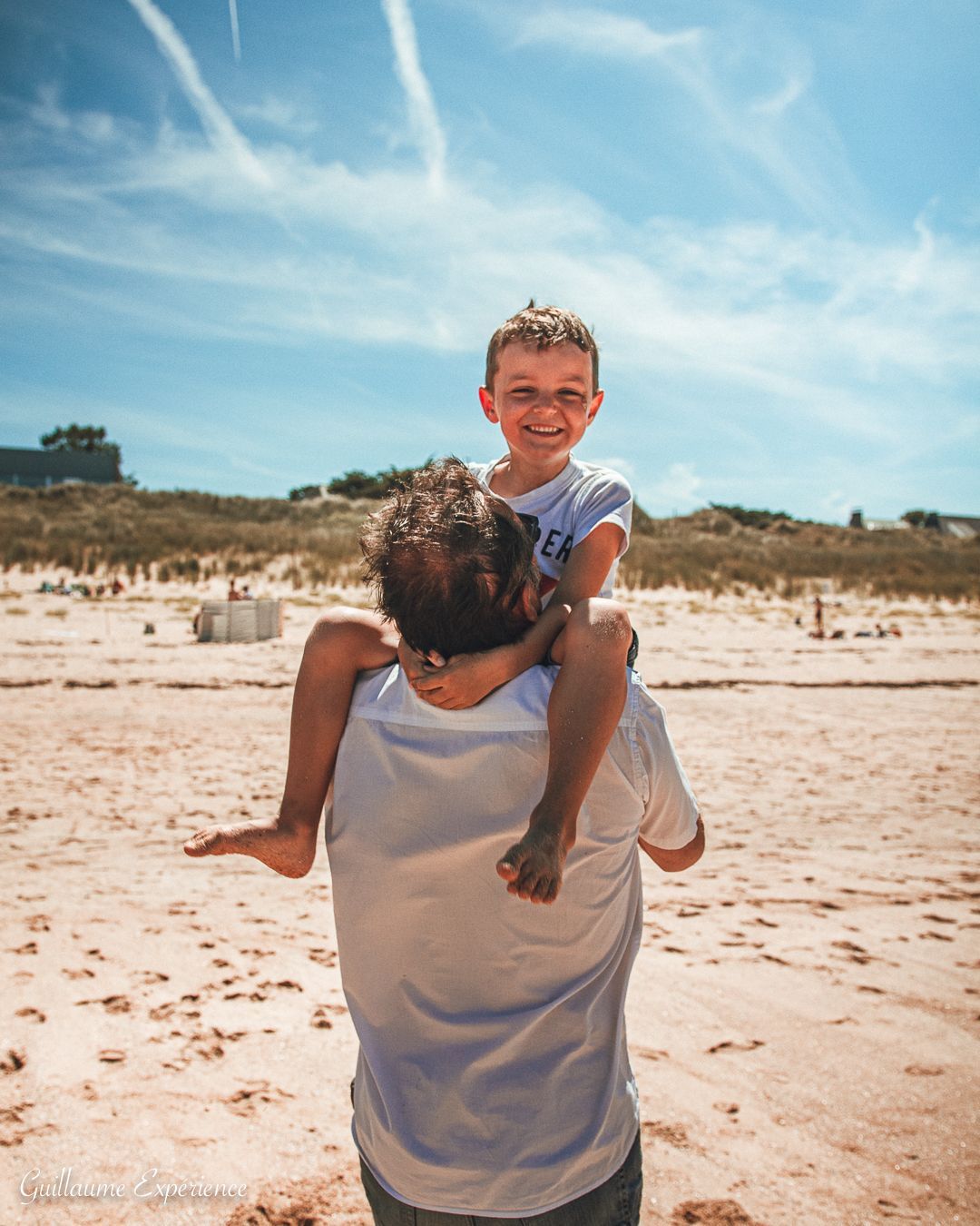 Un père porte un enfant souriant sur ses épaules sur une plage ensoleillée.