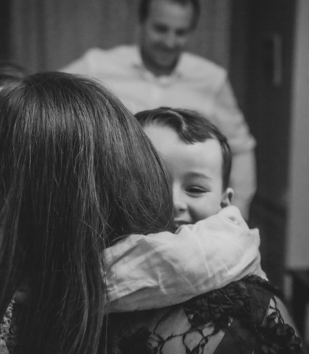 Une femme serre dans ses bras un enfant souriant ; un homme sourit en arrière-plan, le tout en noir et blanc.