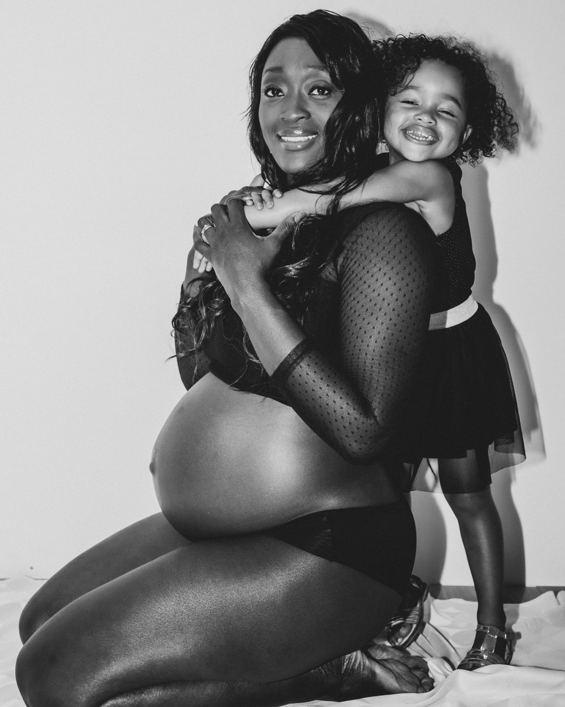 Une femme enceinte, souriante, enlace une jeune fille. Toutes deux sont vêtues de noir, sur fond blanc.