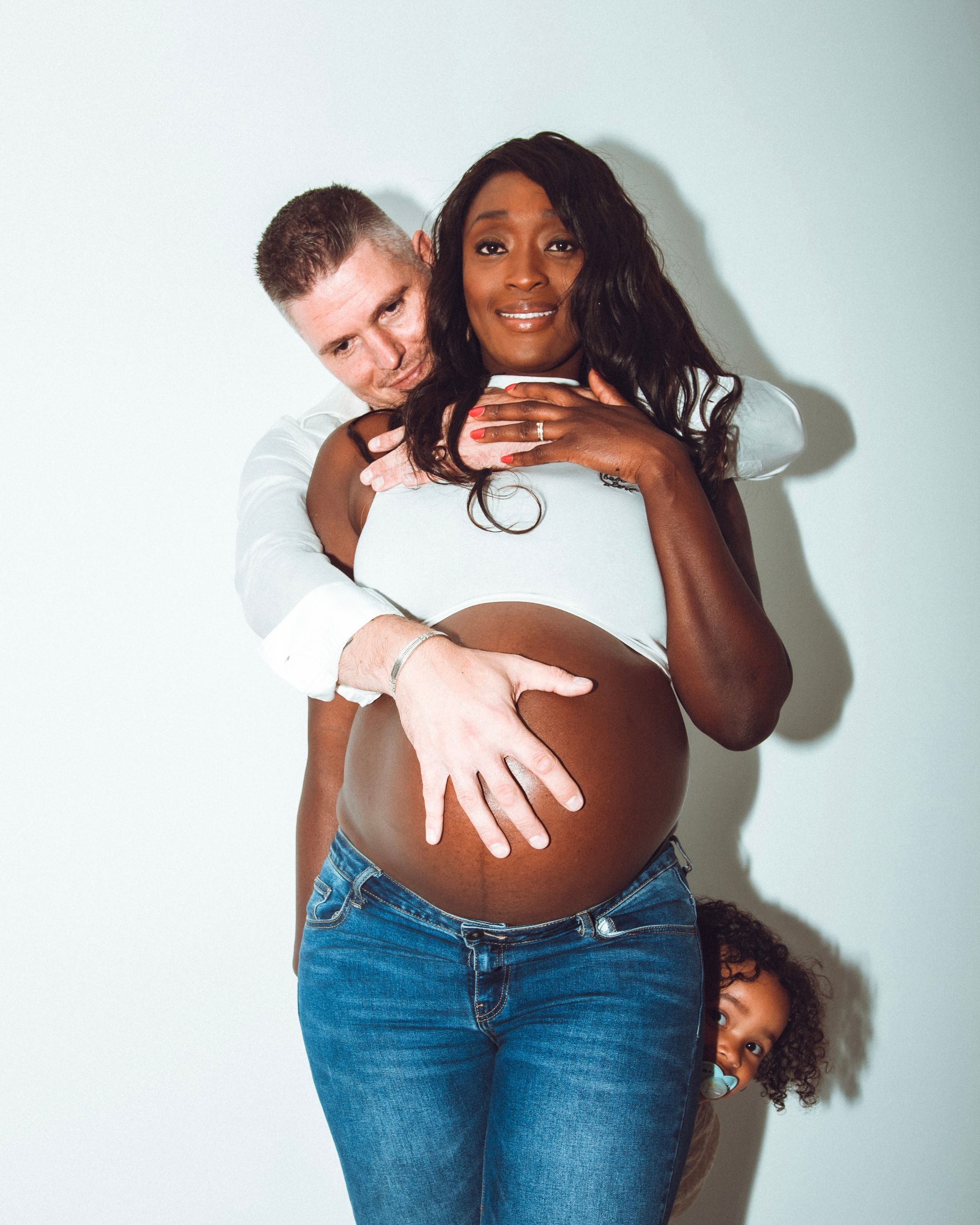 Femme noire enceinte, son partenaire la serrant dans ses bras, son enfant la regardant. Haut blanc, jean, fond blanc.