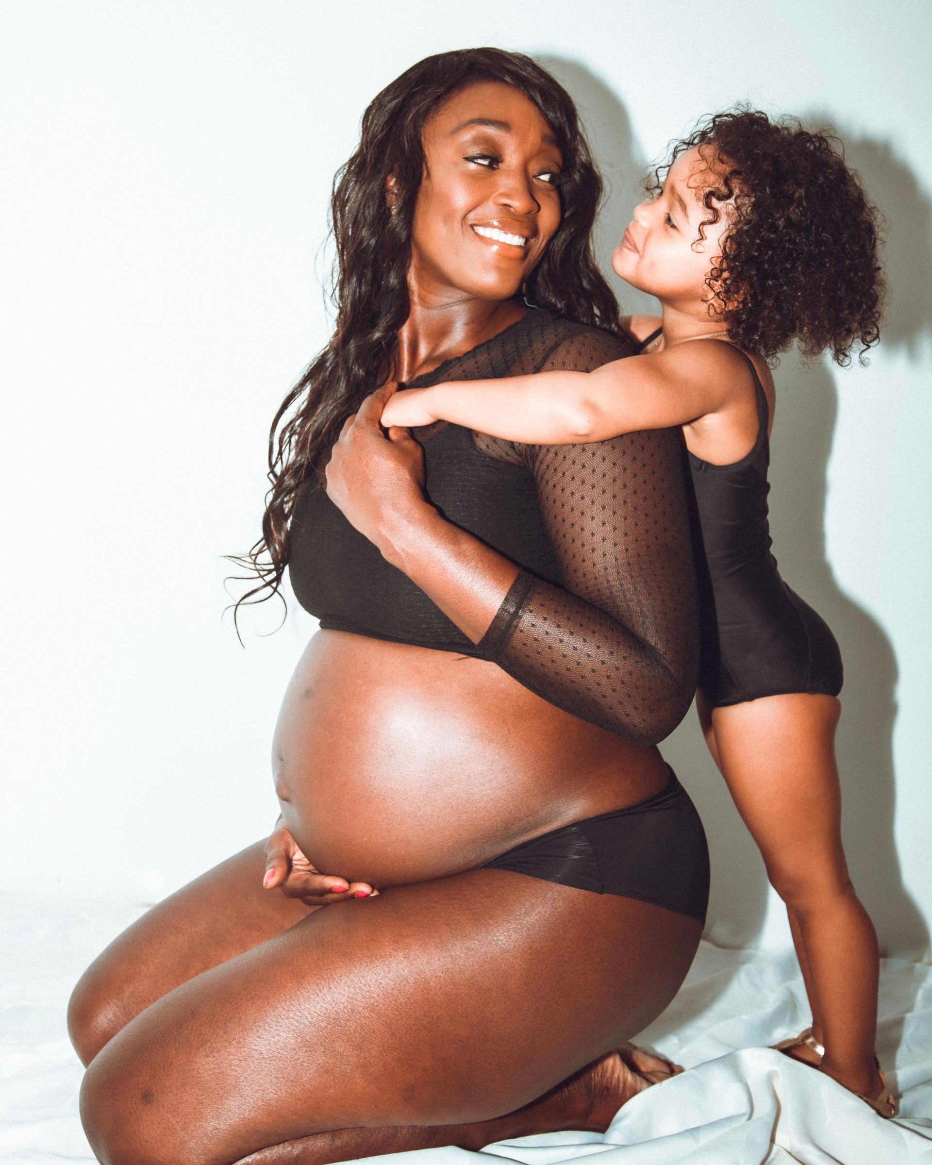 Une femme noire enceinte s'agenouille, souriante, tenant son ventre. Un jeune enfant la serre dans ses bras, levant les .