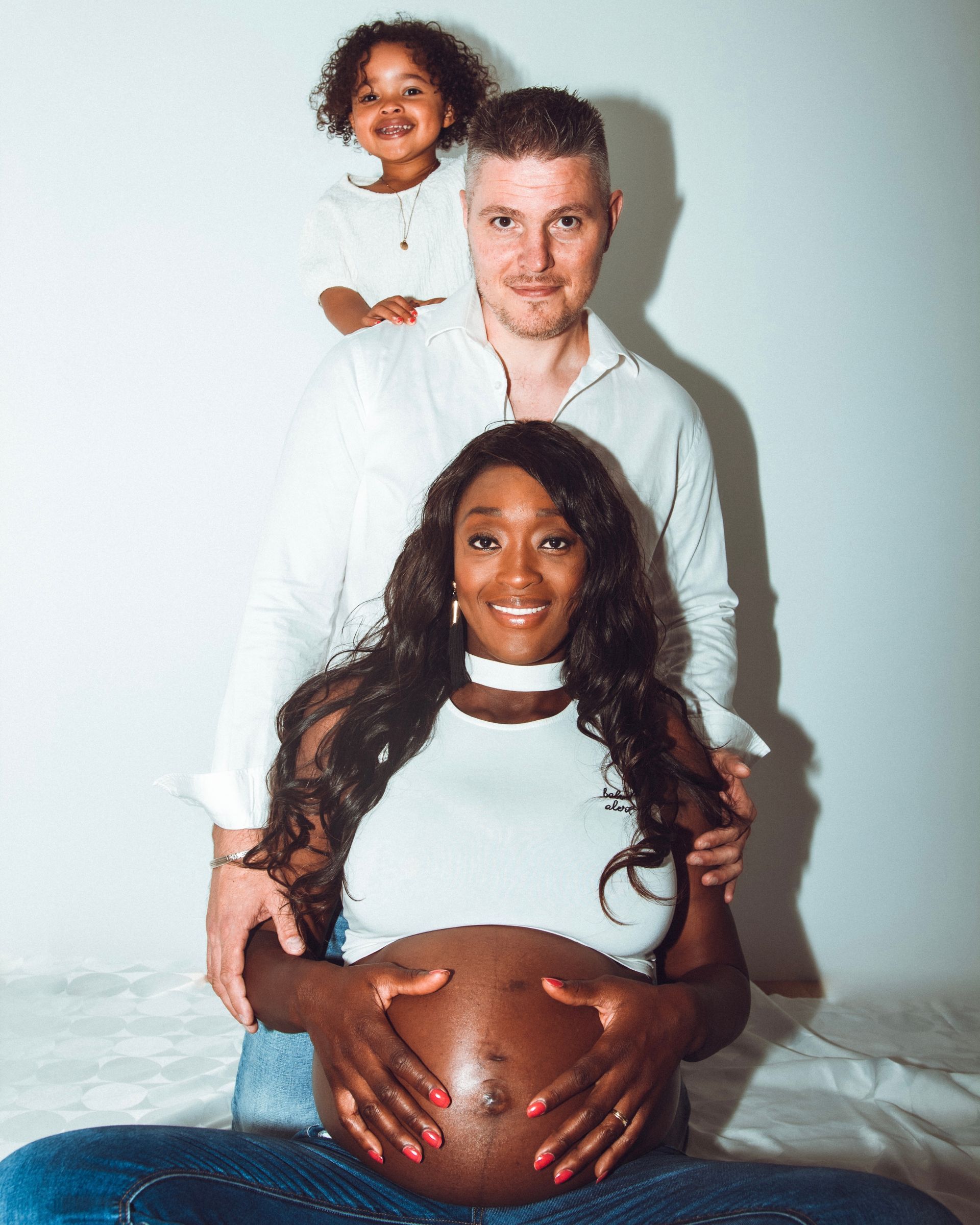 Femme noire enceinte et famille souriante en tenues blanches, à l'intérieur.
