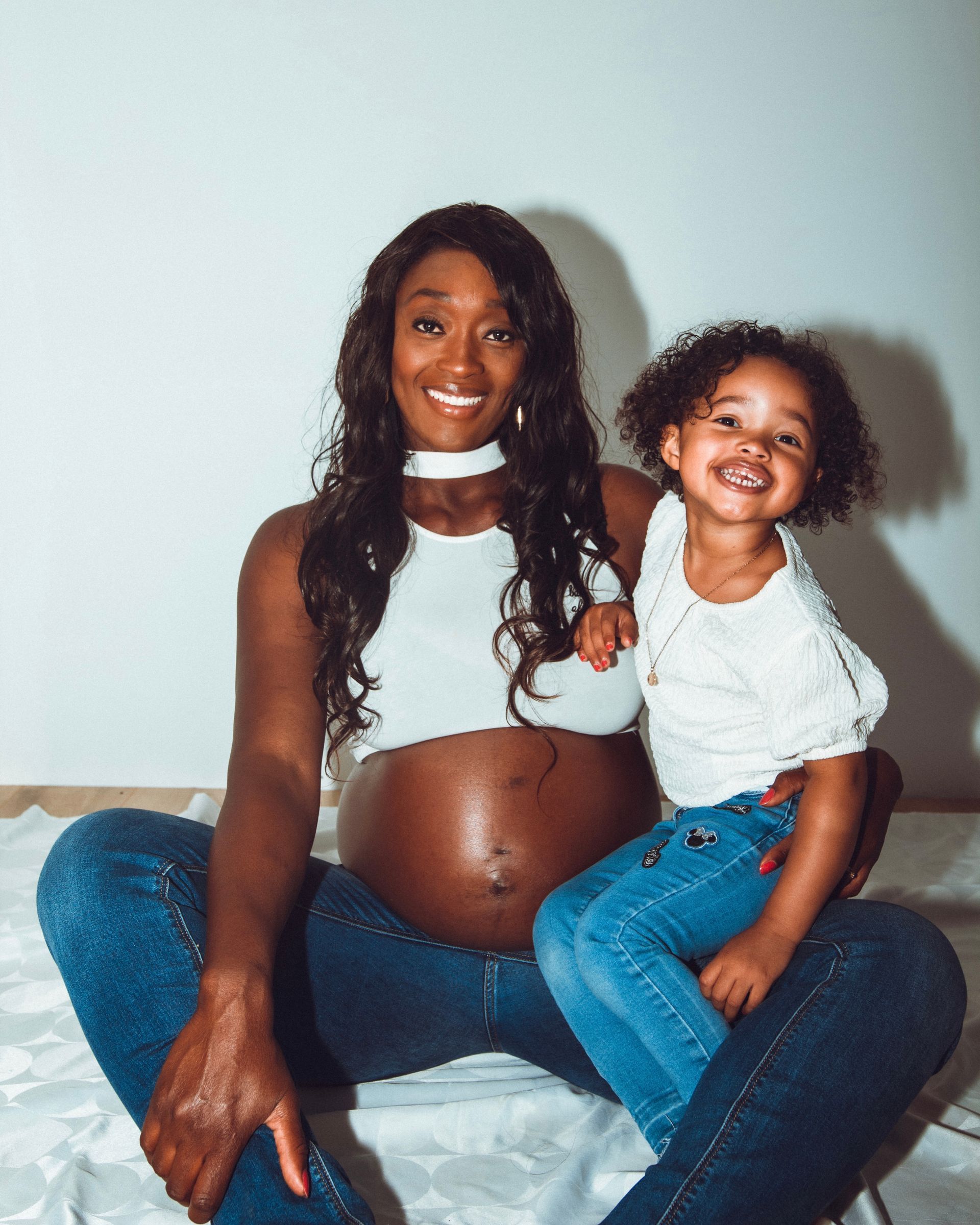 Une femme noire enceinte et un jeune enfant sourient, assis contre un mur blanc.