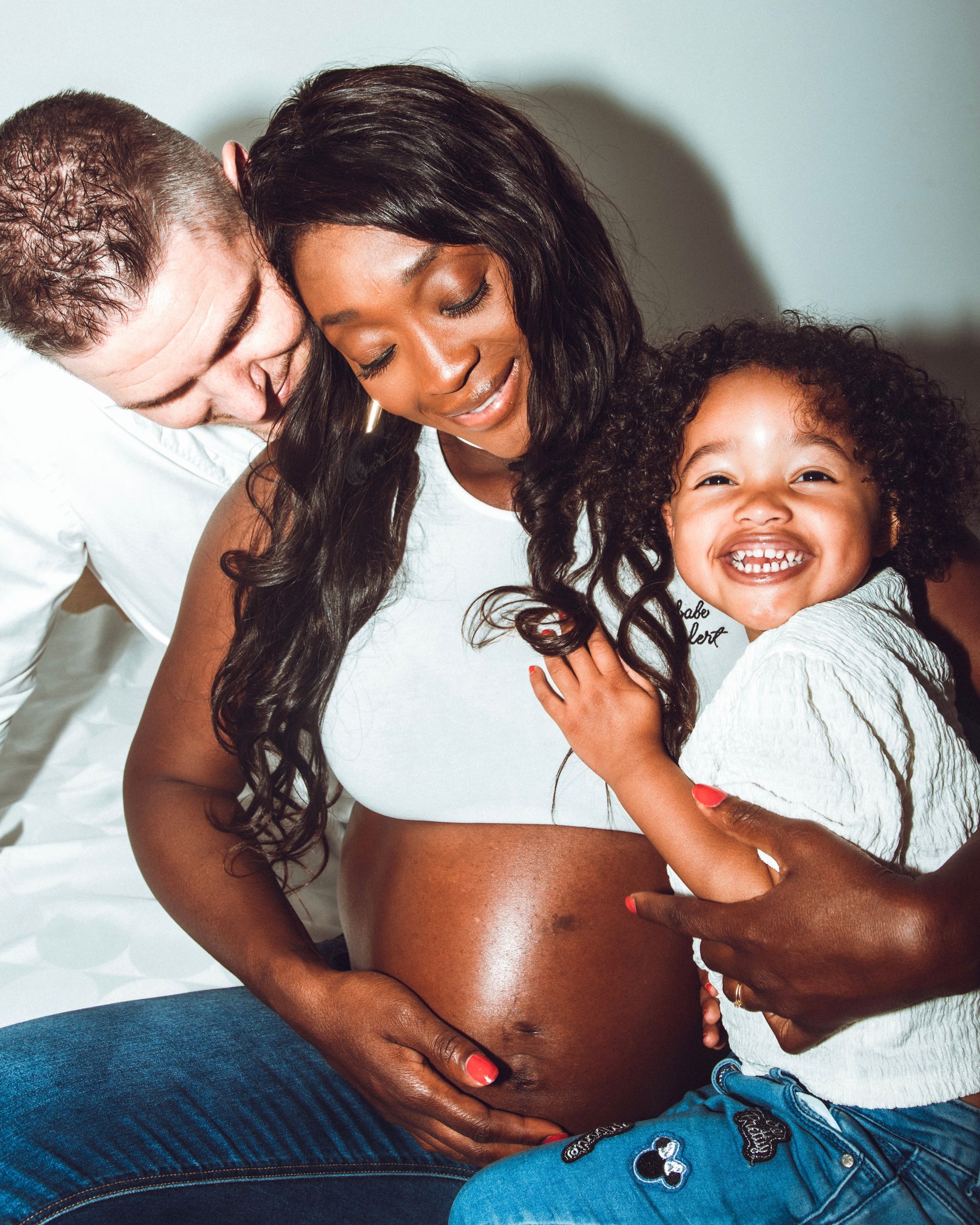 Femme noire enceinte avec son mari et son enfant, souriant, tenant son ventre rond.