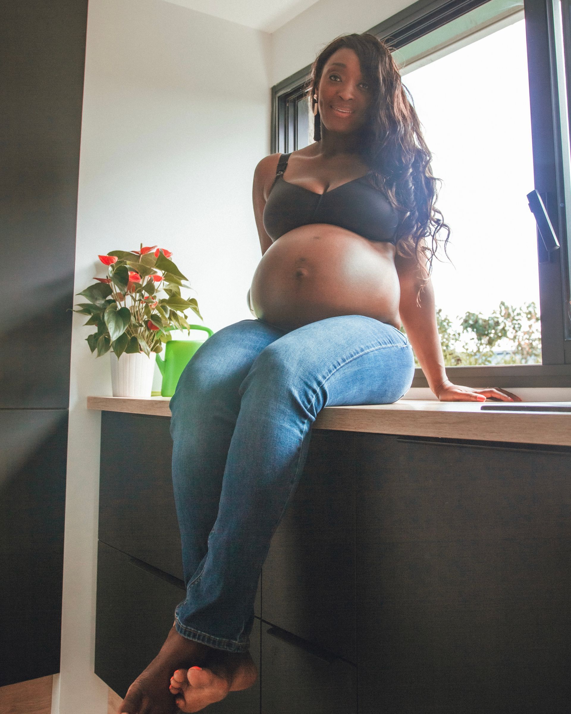 Femme enceinte en soutien-gorge et jean, assise sur un comptoir, regardant la caméra, souriante.