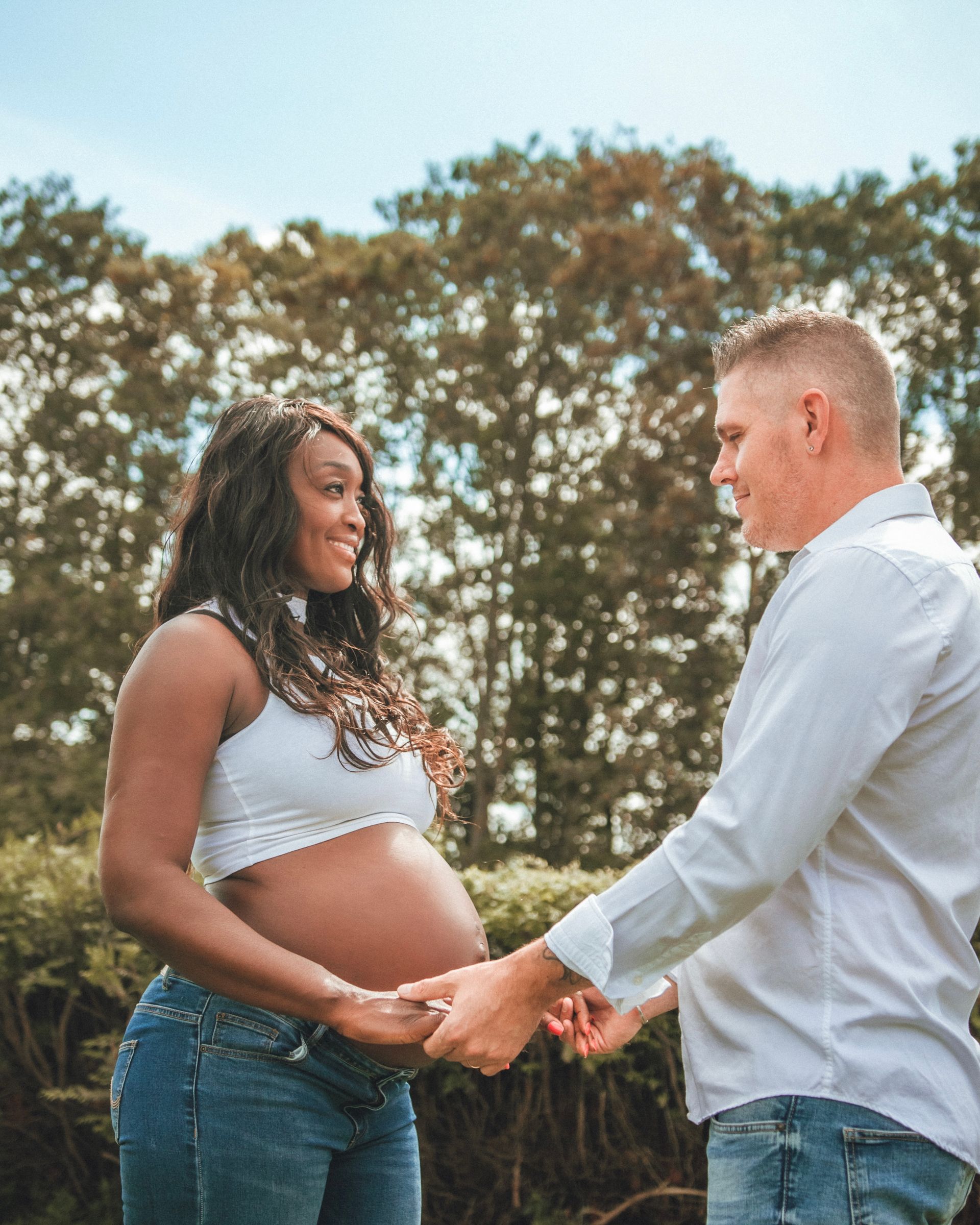 Une femme enceinte et un homme se tenant la main, souriants. En extérieur, sous les arbres.