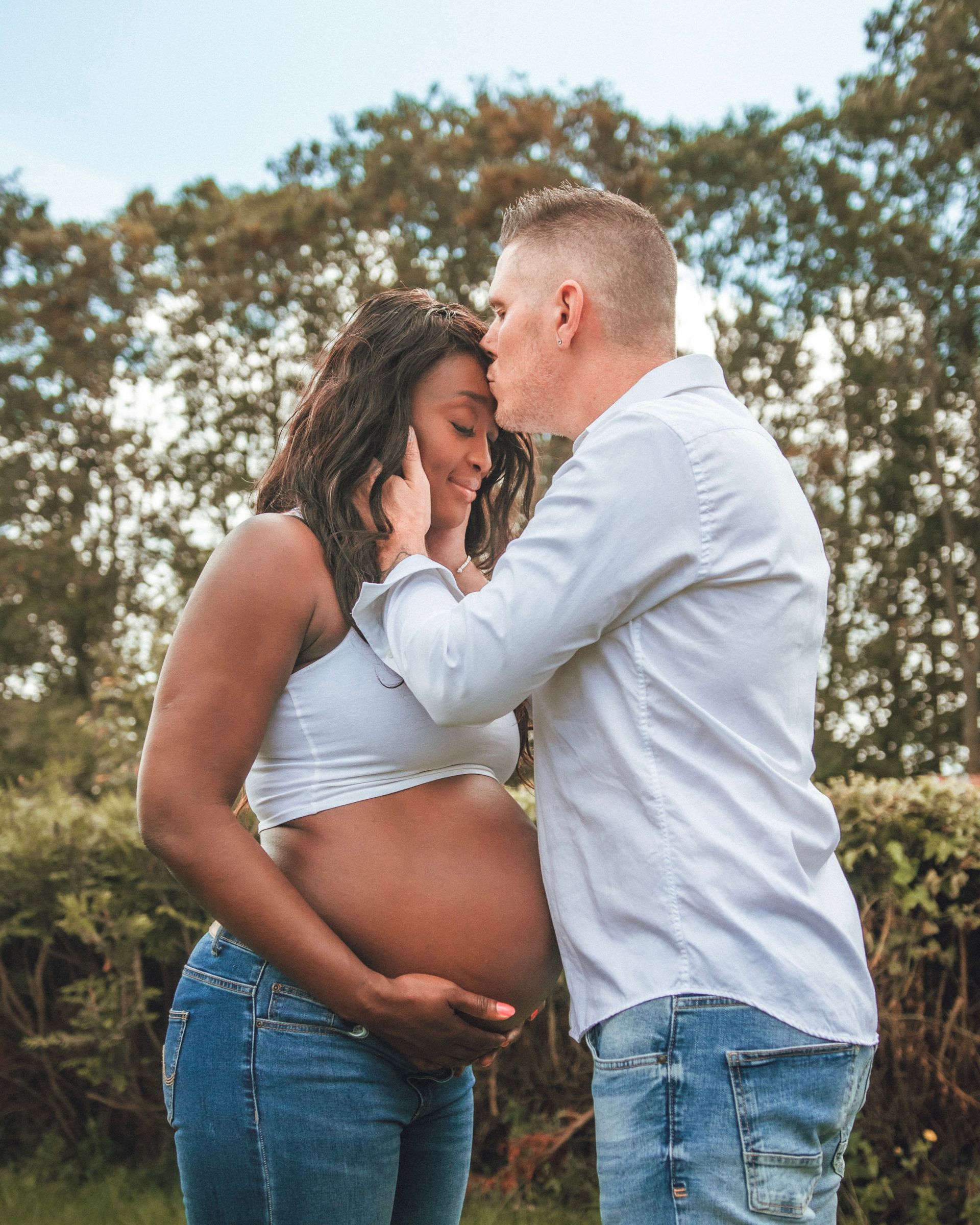 Une femme enceinte avec un homme qui l'embrasse sur le front. Ils sont dehors, entourés de verdure.