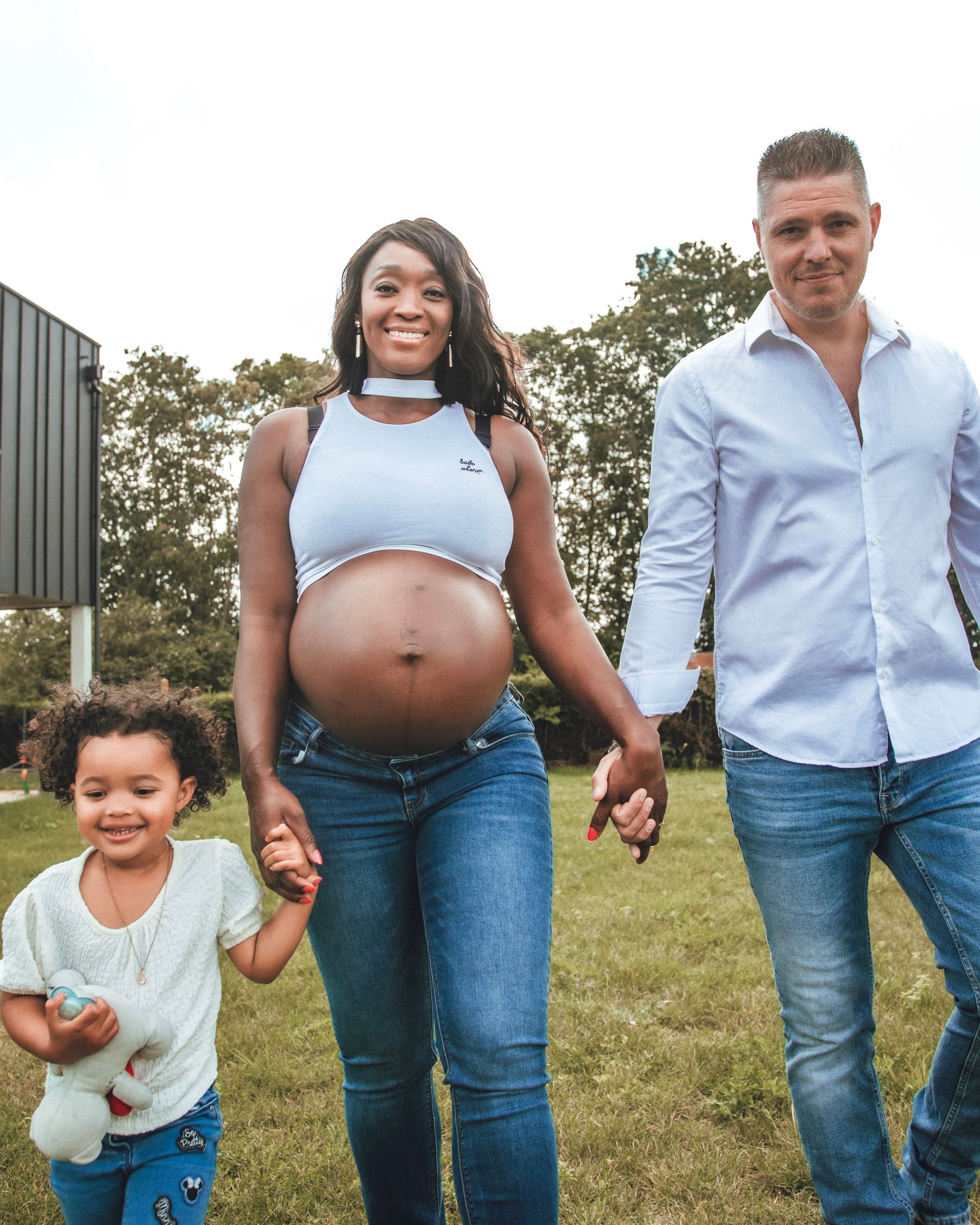 Une femme enceinte, un homme et un enfant marchent dans l'herbe. Ils se tiennent la main et sourient.